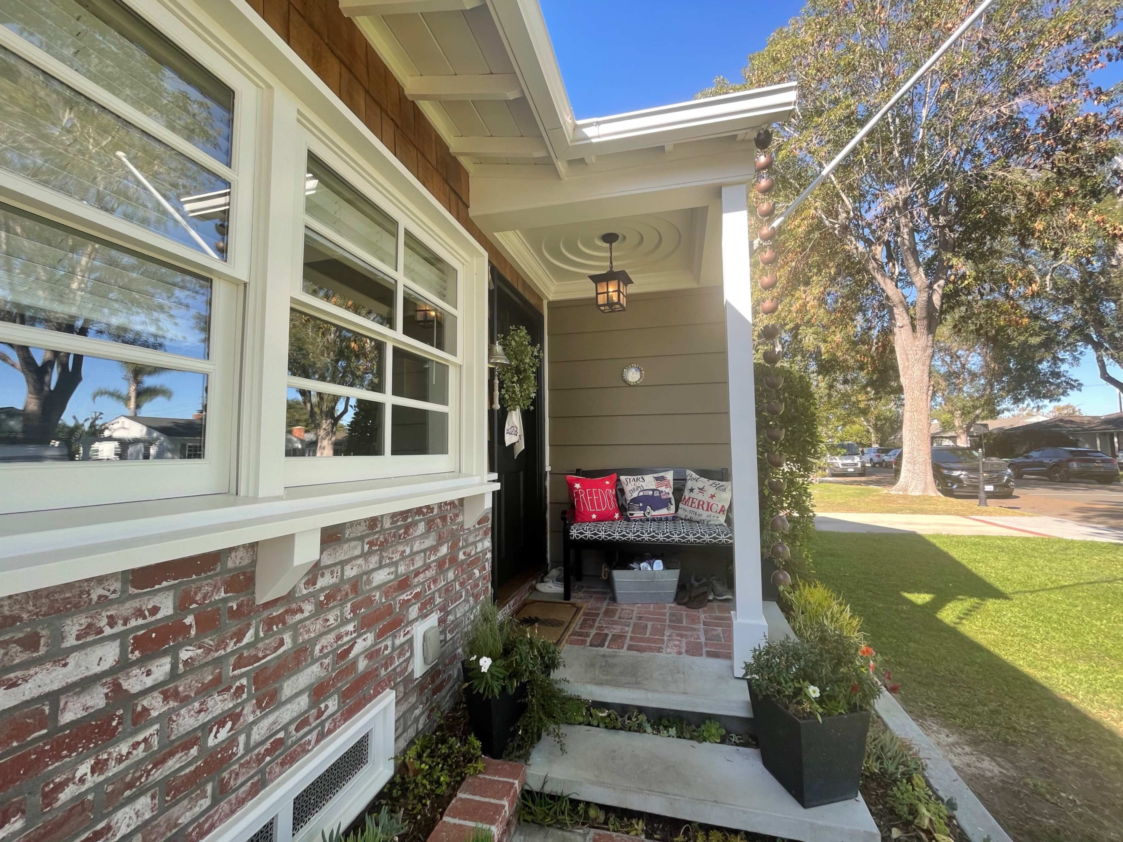 The front porch of a house features a brick wall, a bench with decorative pillows, and a lantern hanging from the ceiling, surrounded by greenery and a pathway leading to the lawn.