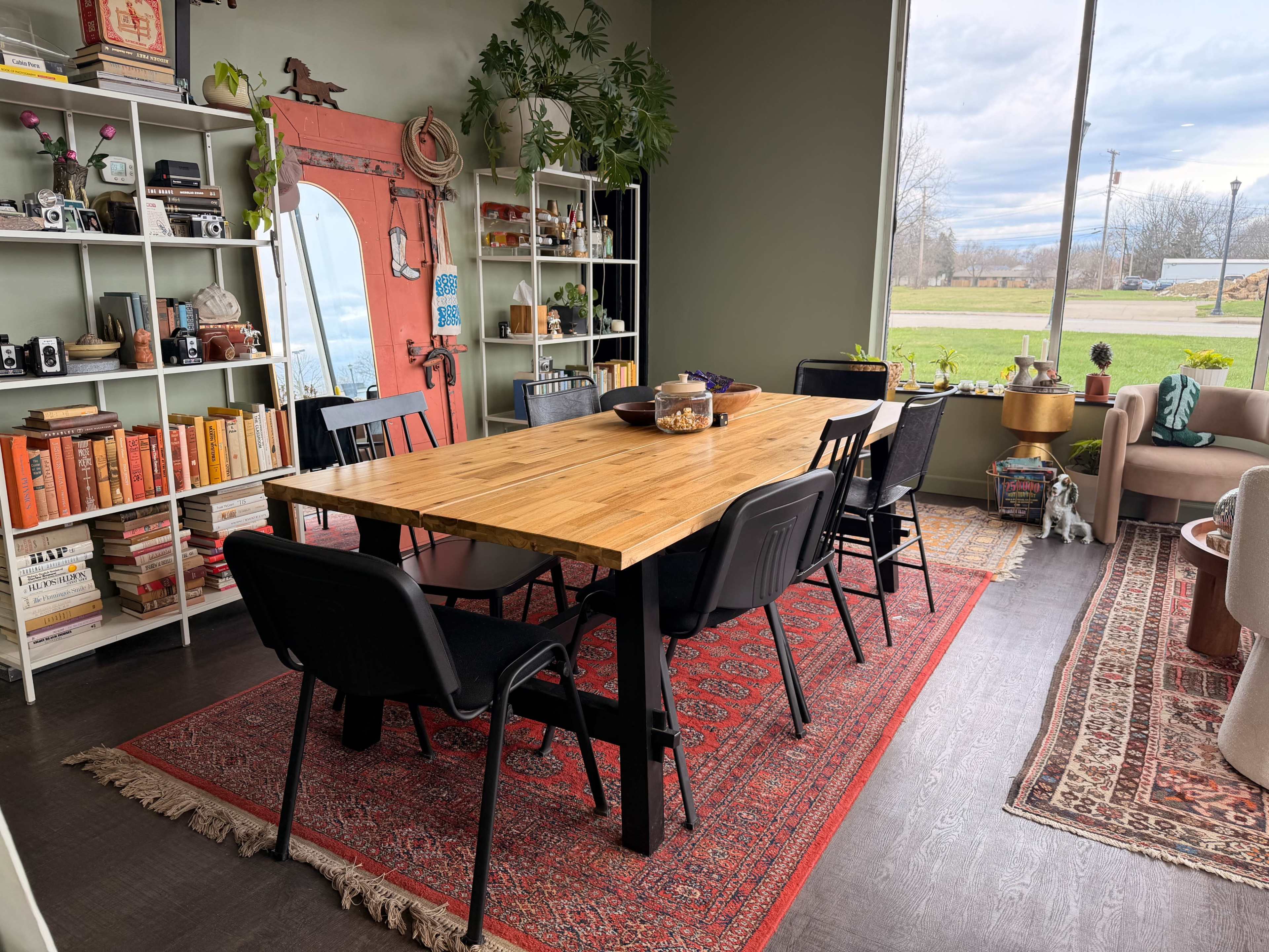 The image shows a spacious dining area with a large wooden table surrounded by black chairs, flanked by shelves filled with books and decorative items, with a window providing a view of a grassy landscape outside.