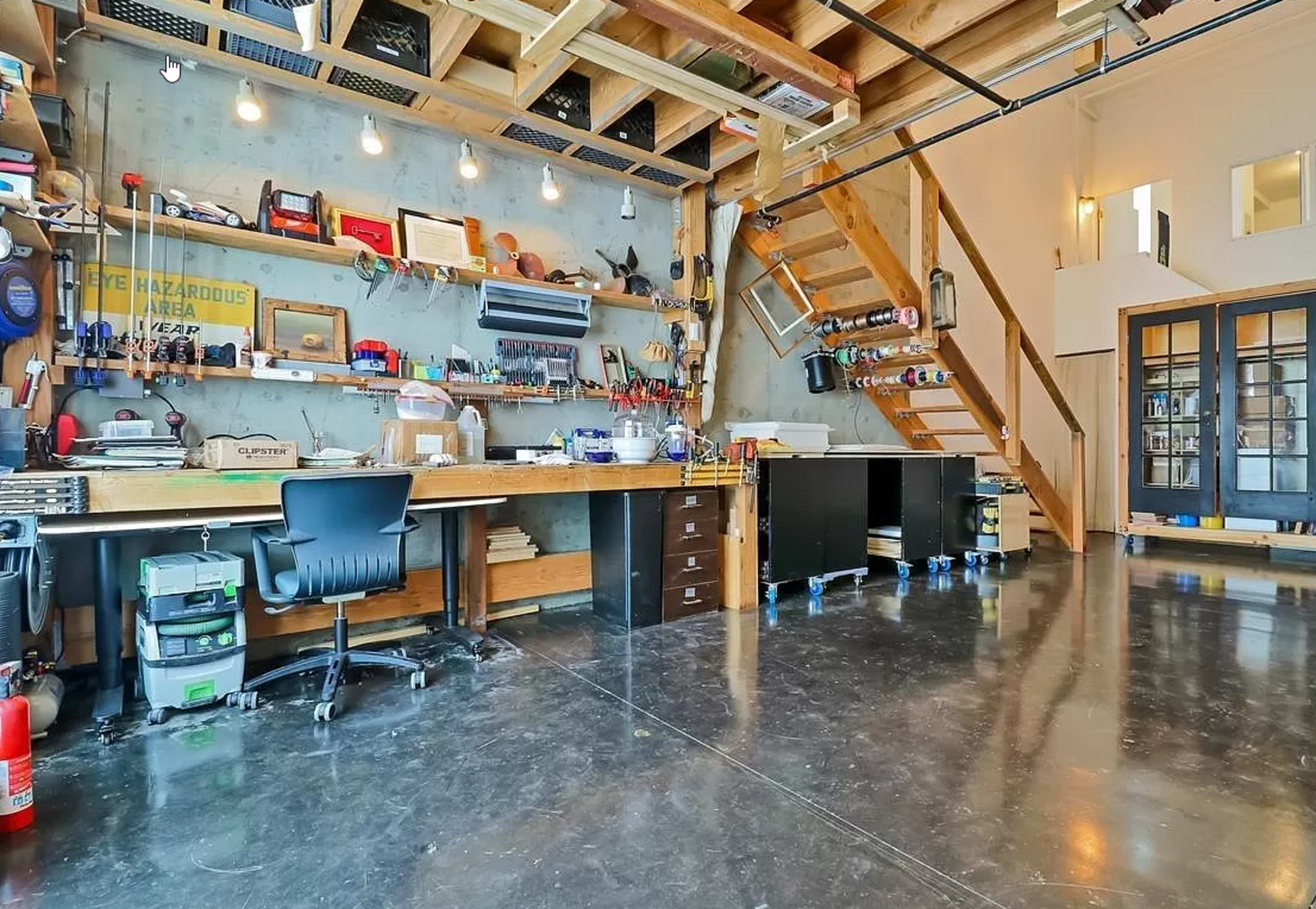 A spacious workshop with a central workbench, shelves filled with tools, and an adjoining staircase leading to a second level.