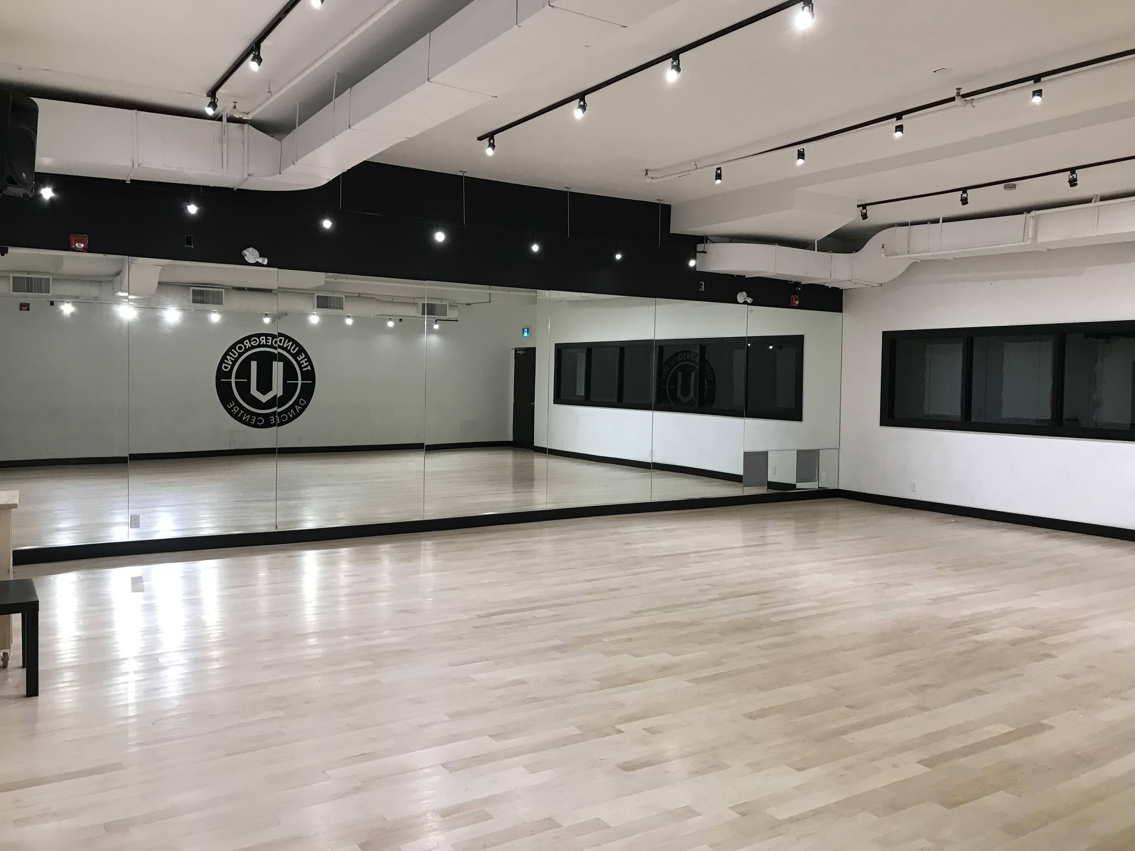 The image shows a spacious dance studio with a wooden floor, large mirrors on one wall, and black and white decor.