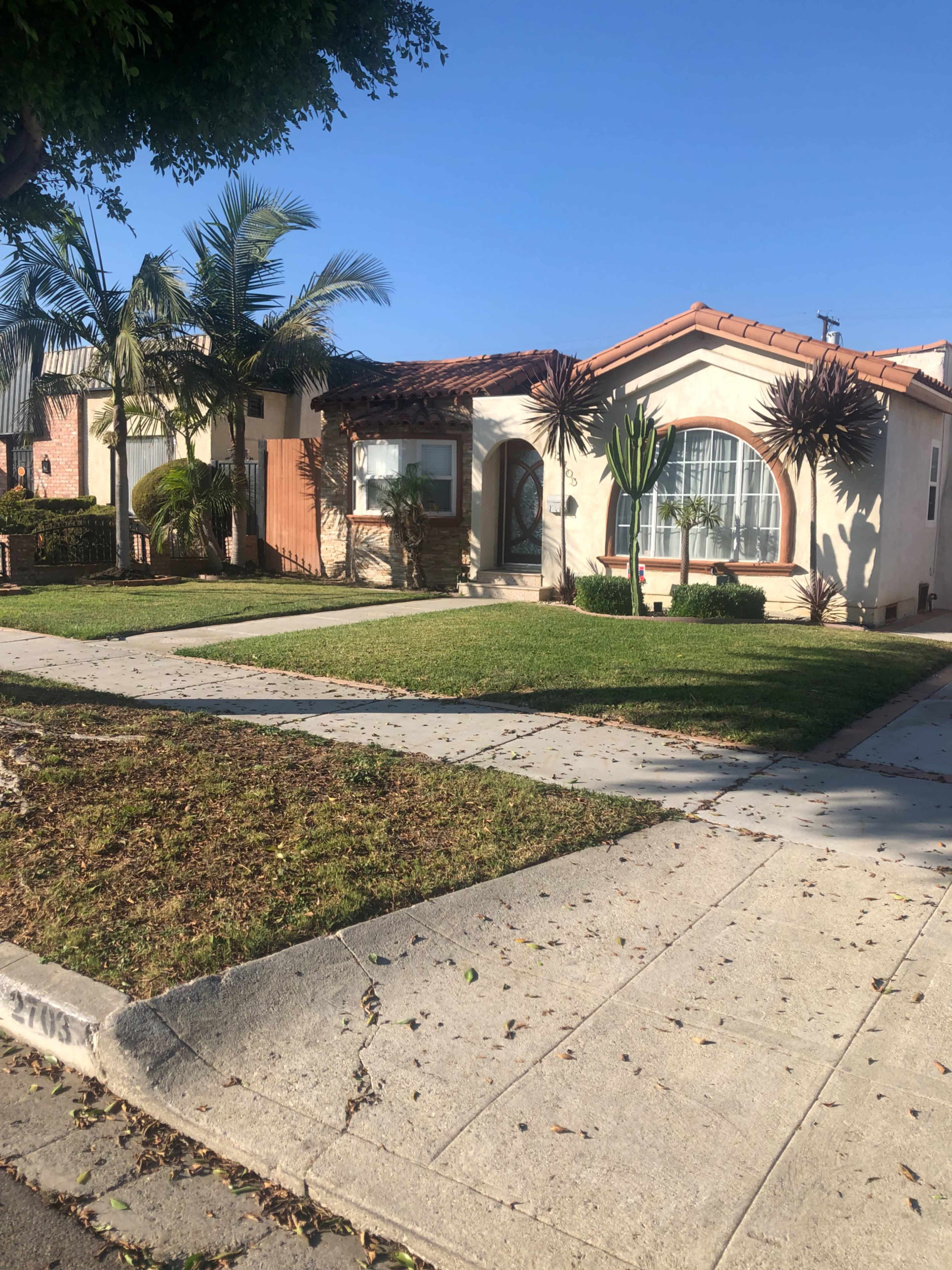 A single-story house with a red-tiled roof and palm trees is located on a grassy corner lot.