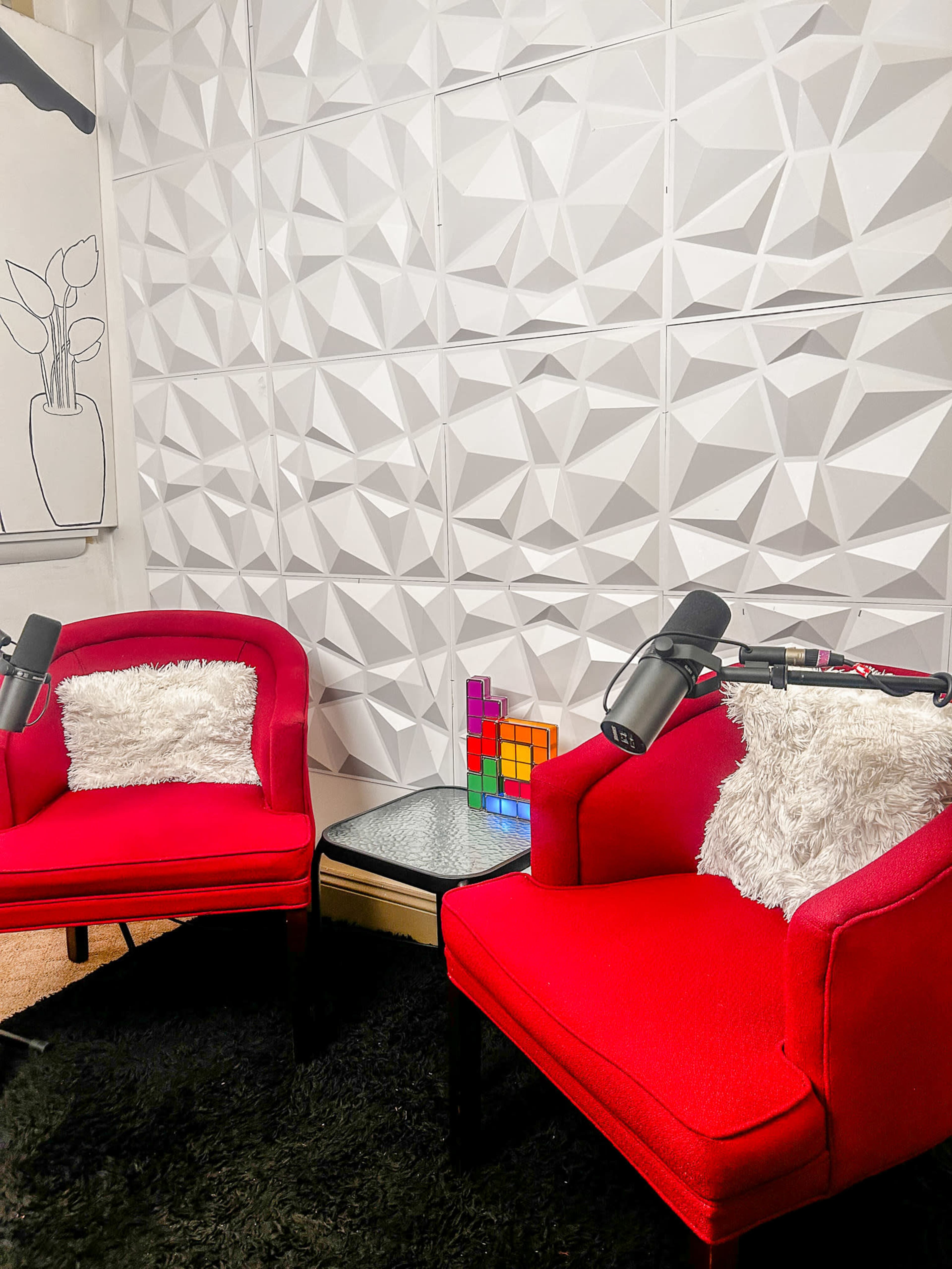 The image shows two red chairs with white pillows facing each other, positioned in front of a textured white wall, with a colorful Tetris-style lamp and two microphones on the chairs.