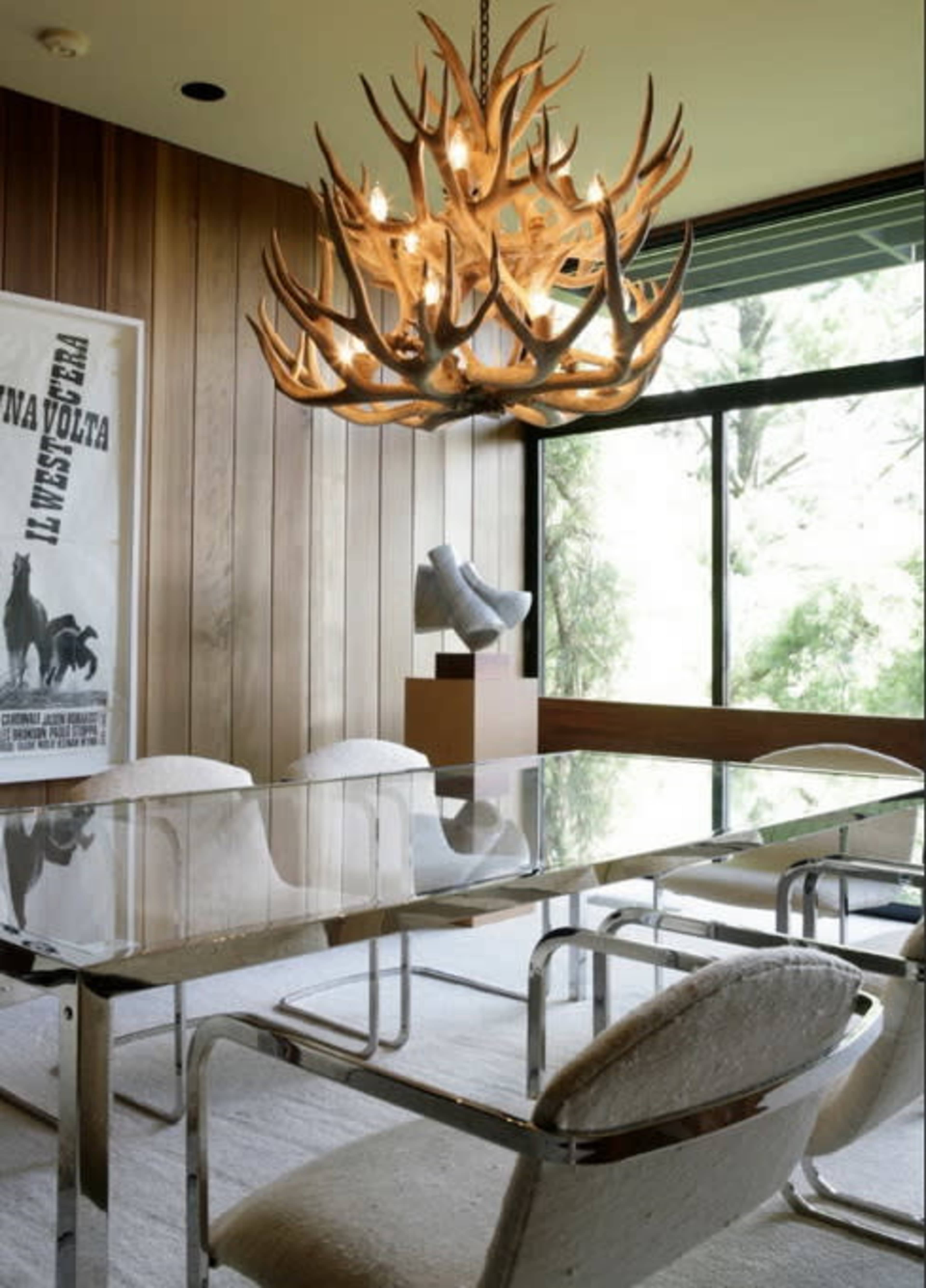 A modern dining room with a glass table, minimalist chairs, and a large antler chandelier hanging from the ceiling, surrounded by wooden paneling and a window view.