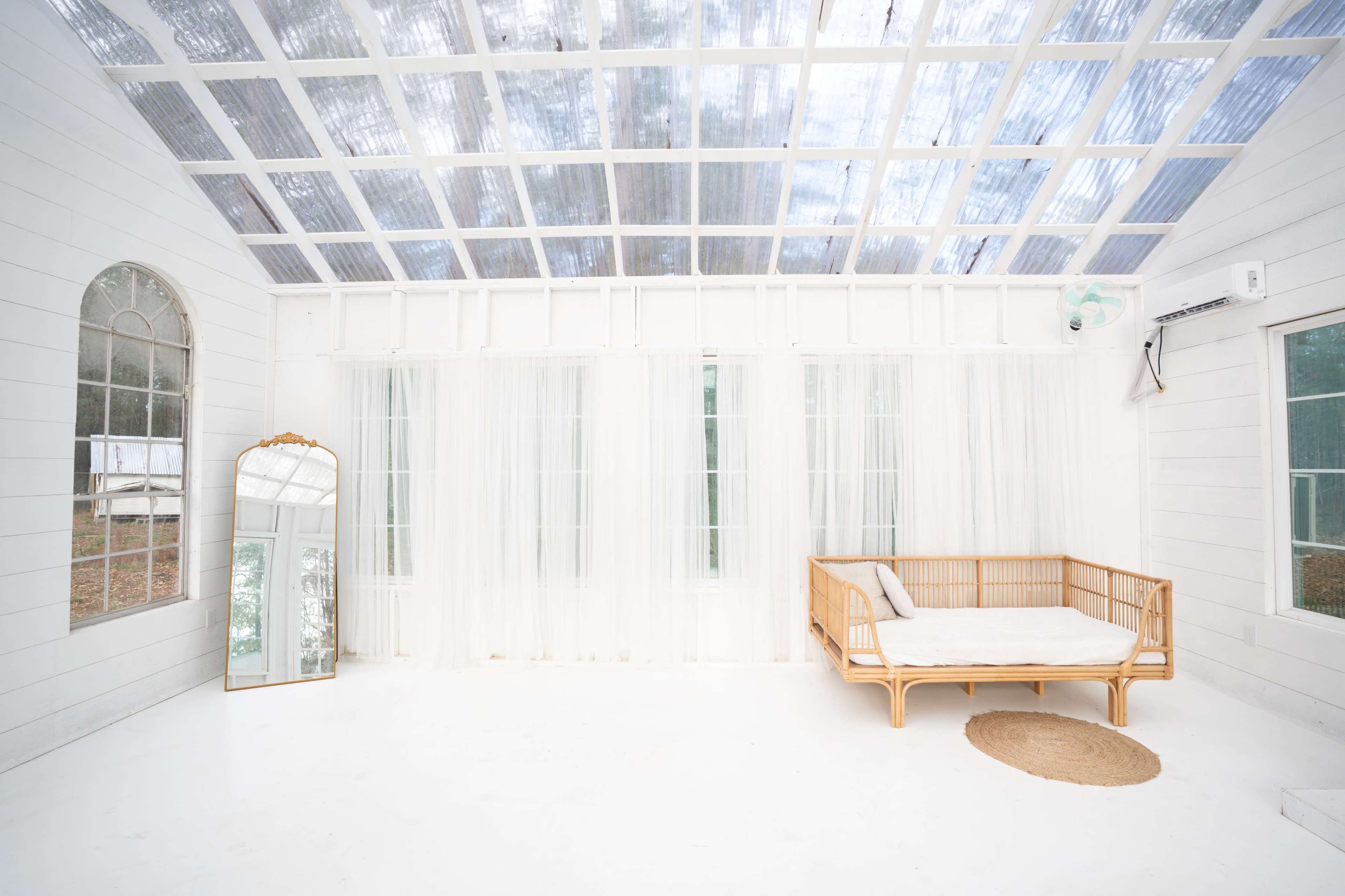 The image shows a bright, airy indoor space with white walls, large windows, a rattan sofa, and a round jute rug on a white floor.