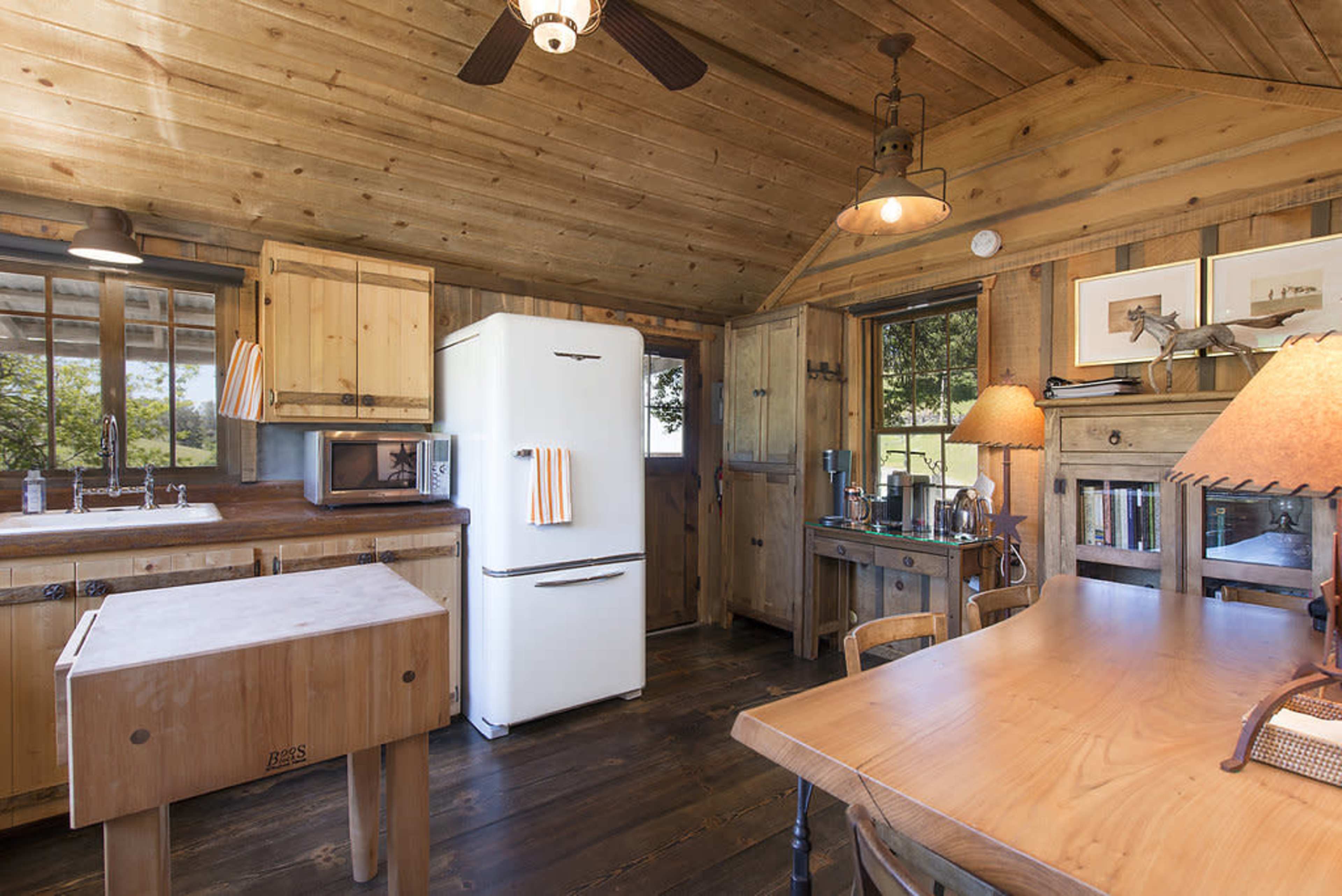 The image shows a wooden cabin kitchen equipped with a refrigerator, a table, and various kitchen elements under warm lighting.