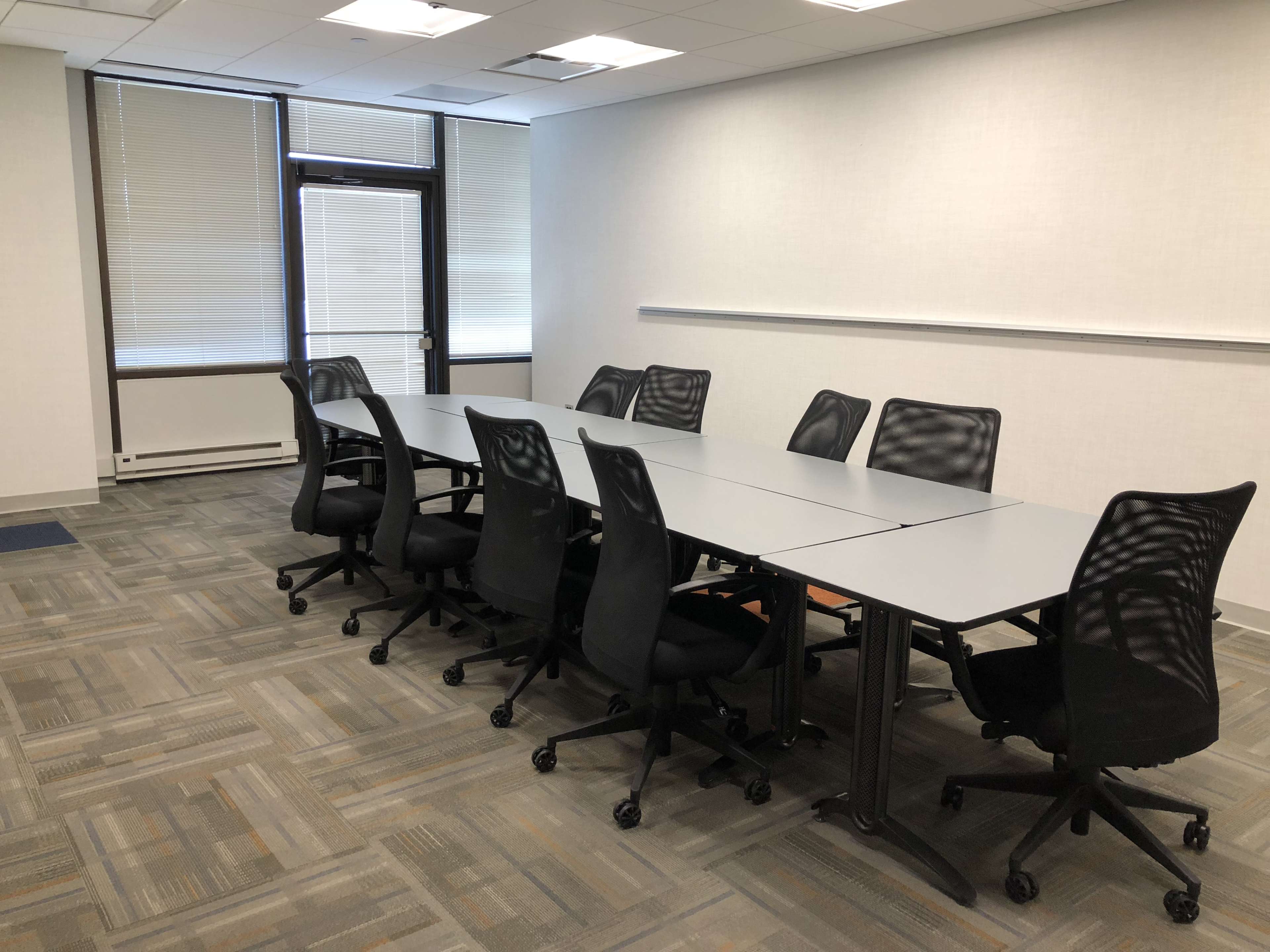 A large, empty conference room features a long table surrounded by black rolling chairs, with windows allowing natural light to enter.