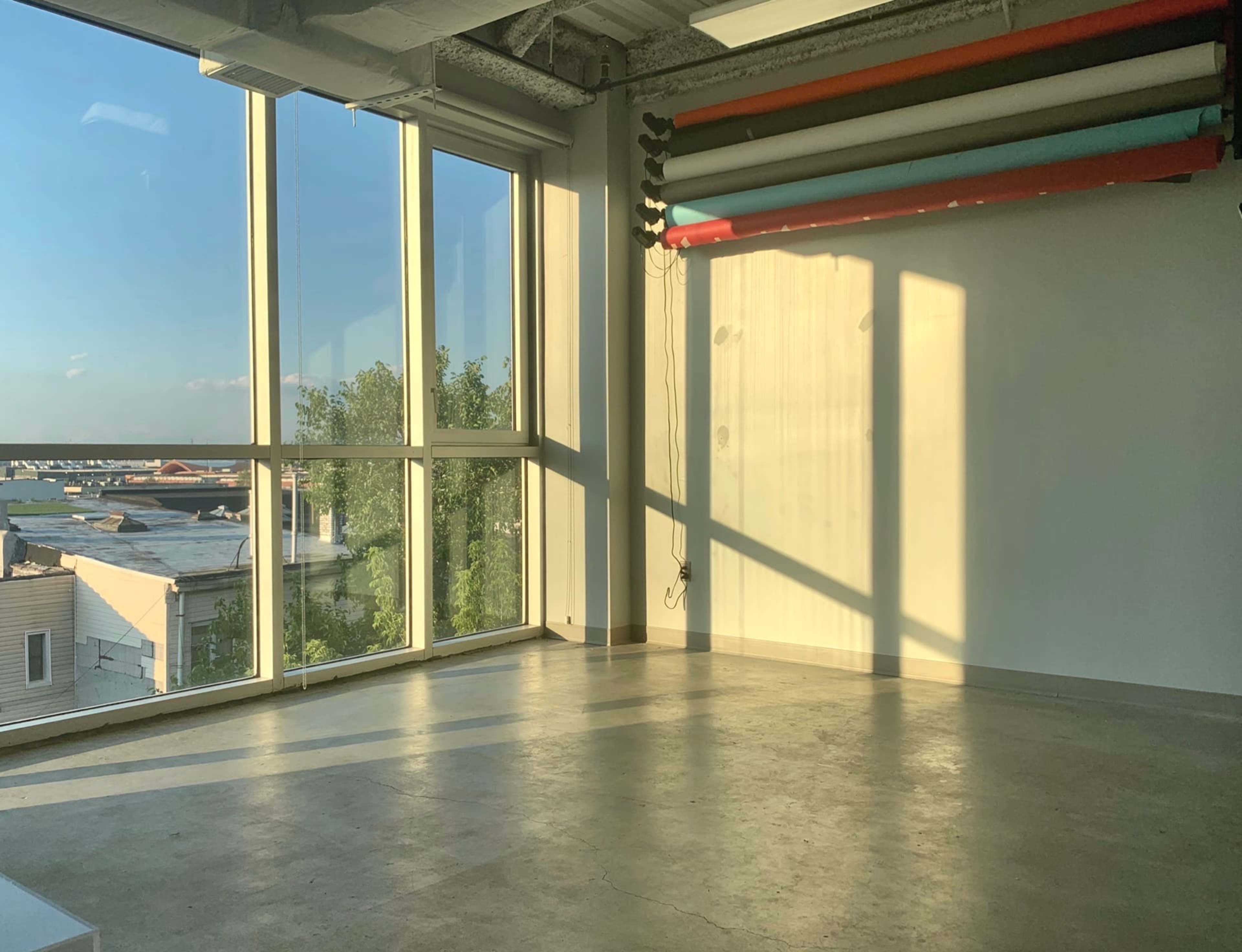 A spacious, empty room with large windows allowing sunlight to cast shadows on the concrete floor.