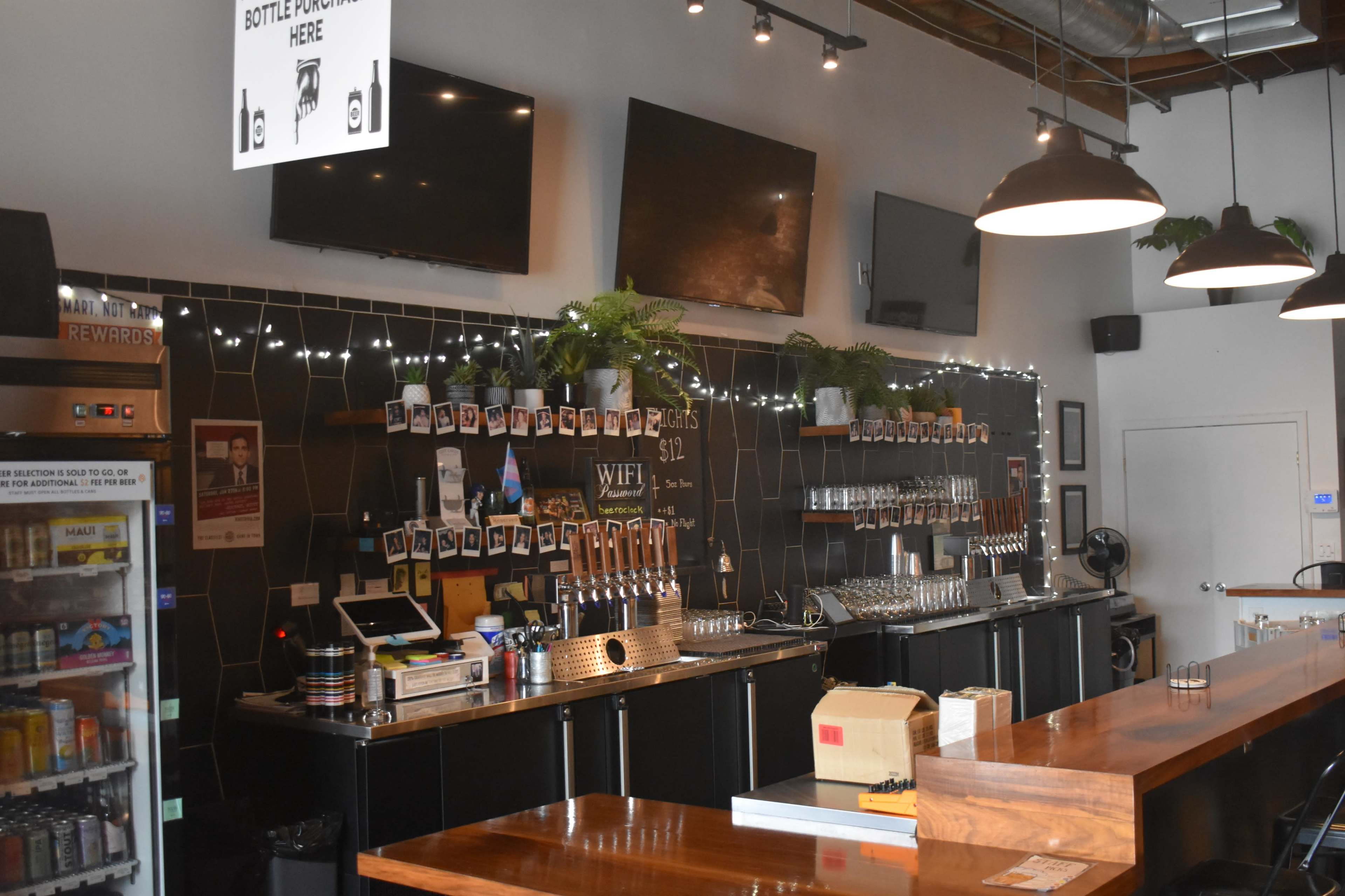 The image shows a bar area with a long wooden counter, multiple beer taps, a wall of framed photos, and several TVs mounted above the bar.