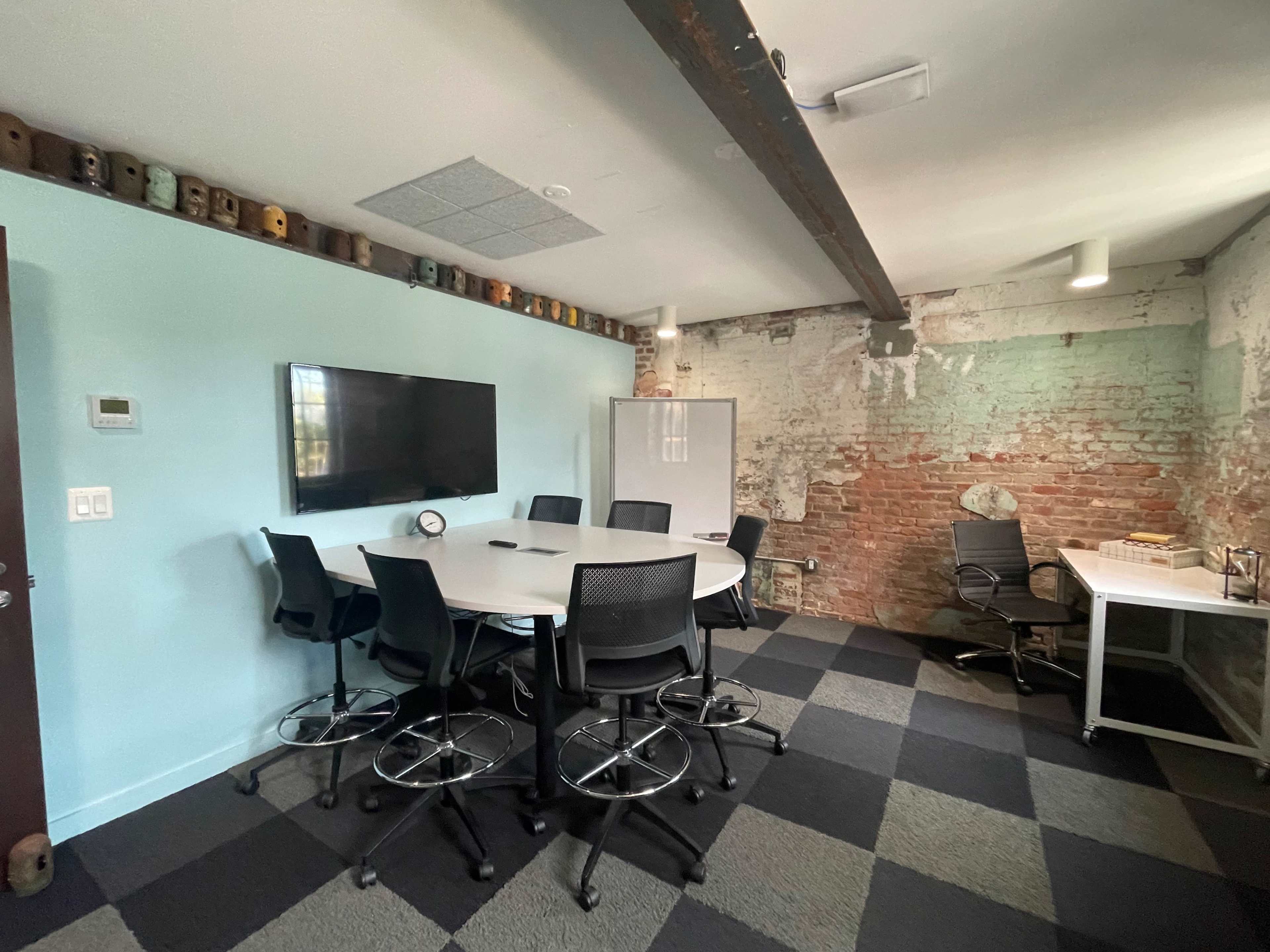 A conference room features a circular table surrounded by black chairs, a large monitor on the wall, and exposed brick walls with a section for an office desk in the corner.