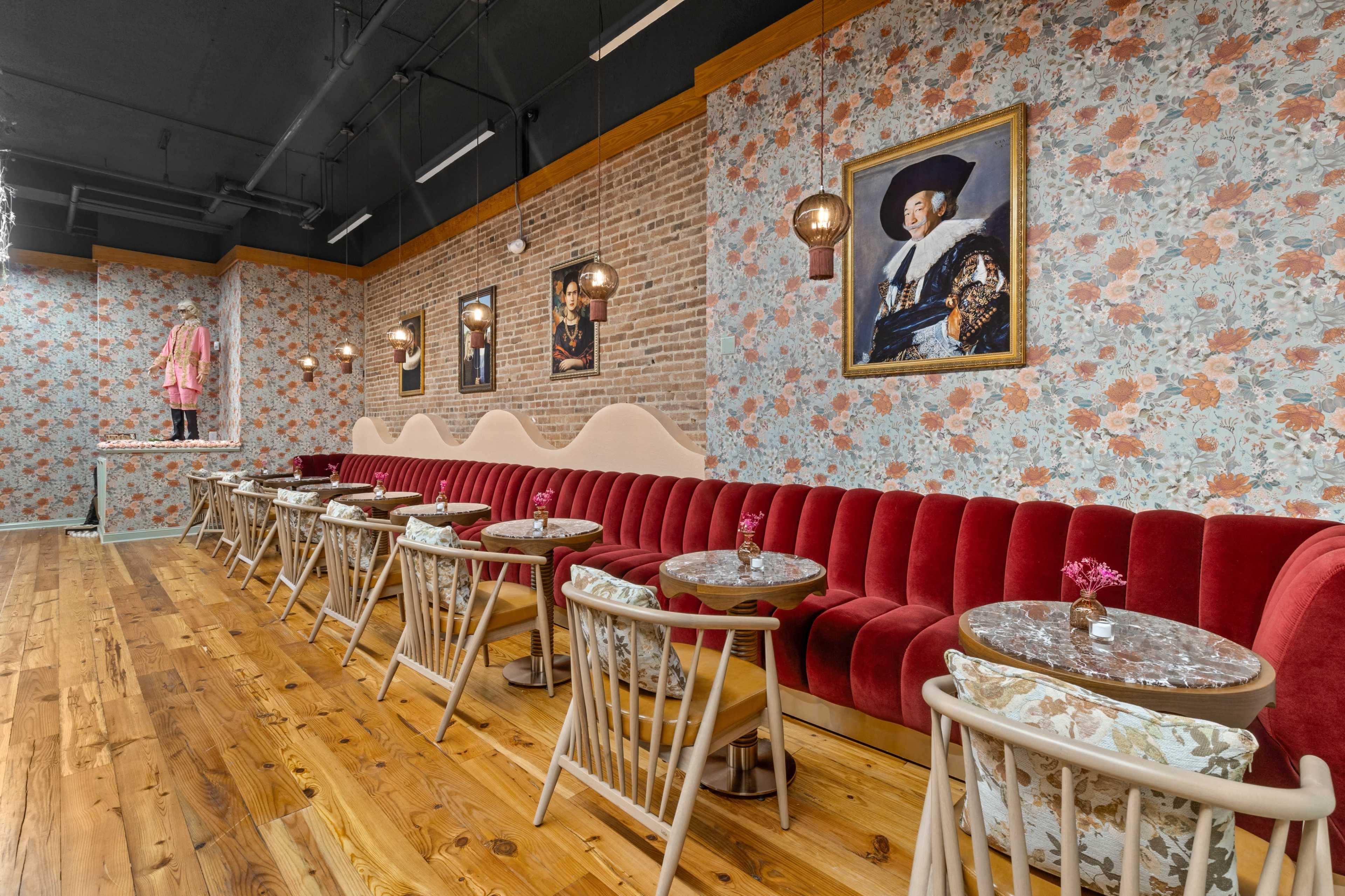 The image shows a decorated café interior with a red velvet bench, floral wallpaper, wooden flooring, and small round tables.