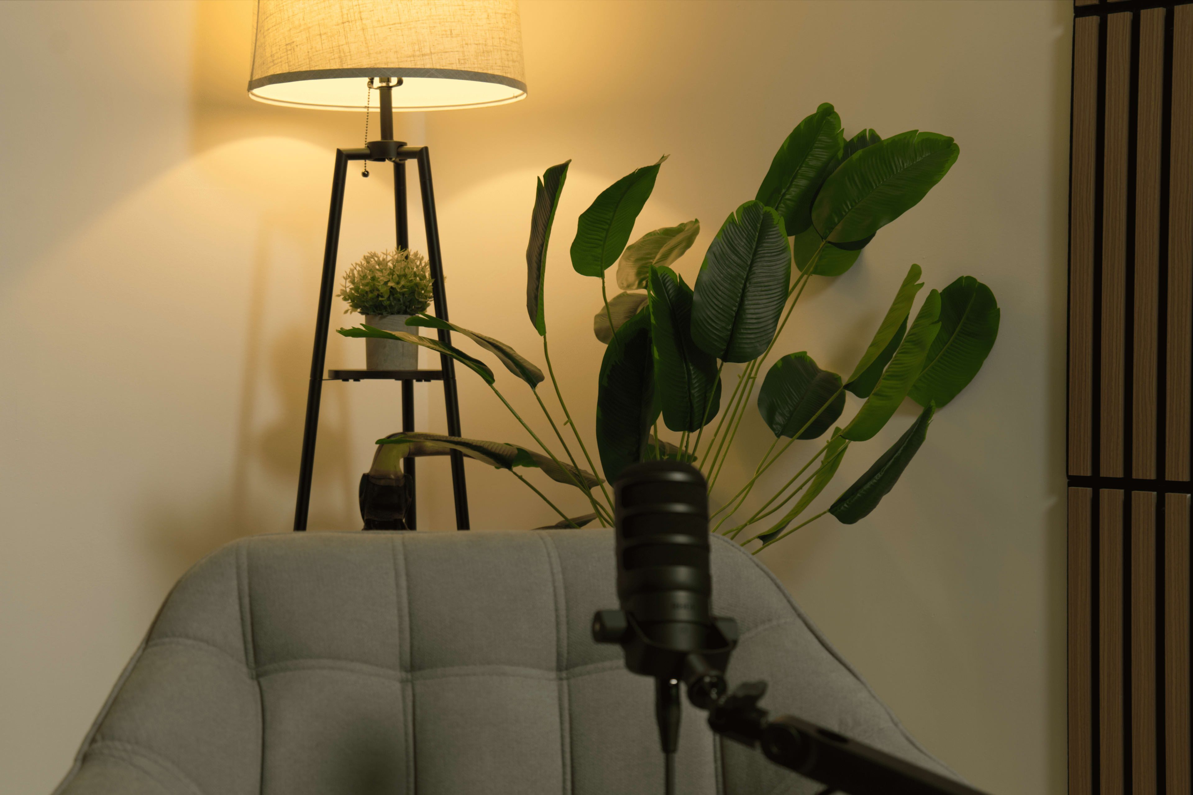 A gray upholstered chair is positioned in front of a potted plant and a tripod lamp emitting soft light against a white wall.