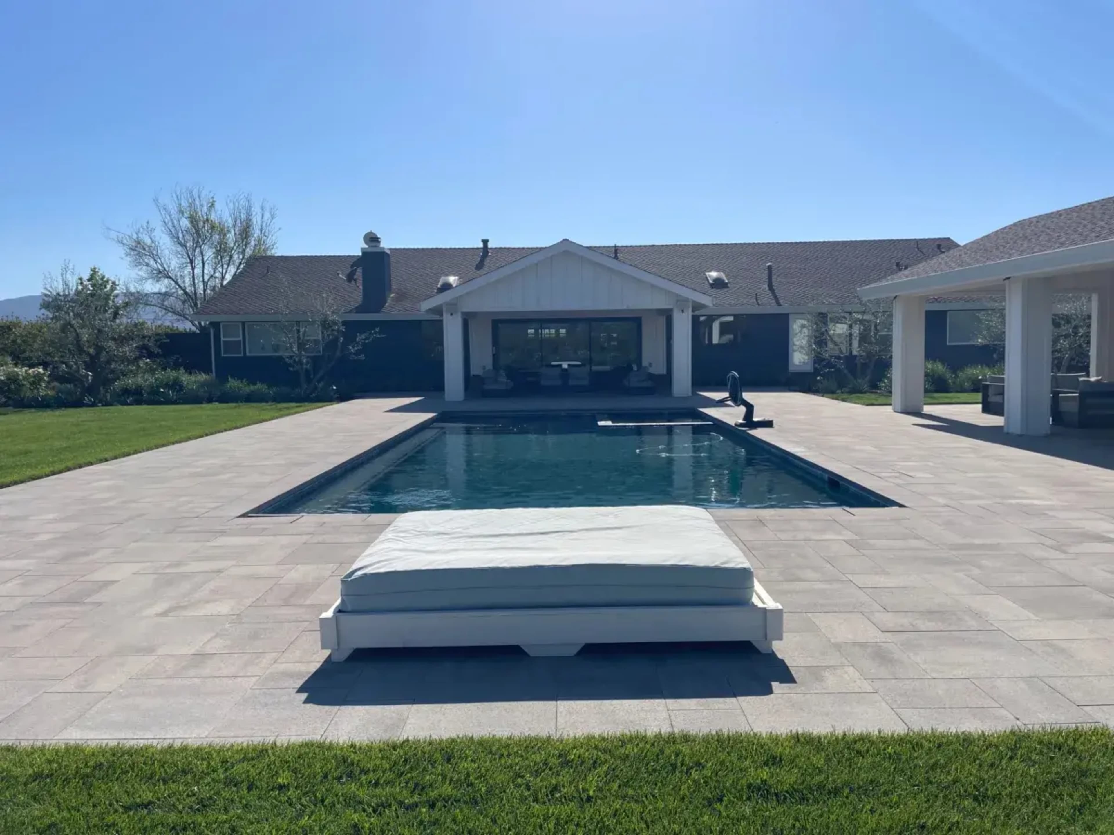 A flat, rectangular swimming pool is located in the center of a patio surrounded by a well-manicured lawn and a house in the background.