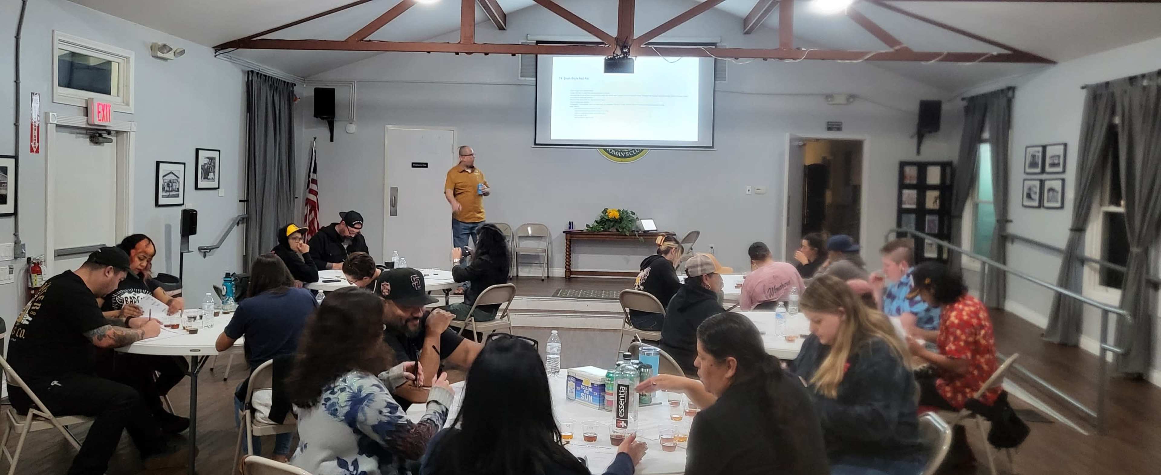 A group of people is seated at tables in a hall, with a speaker presenting on a screen at the front of the room.