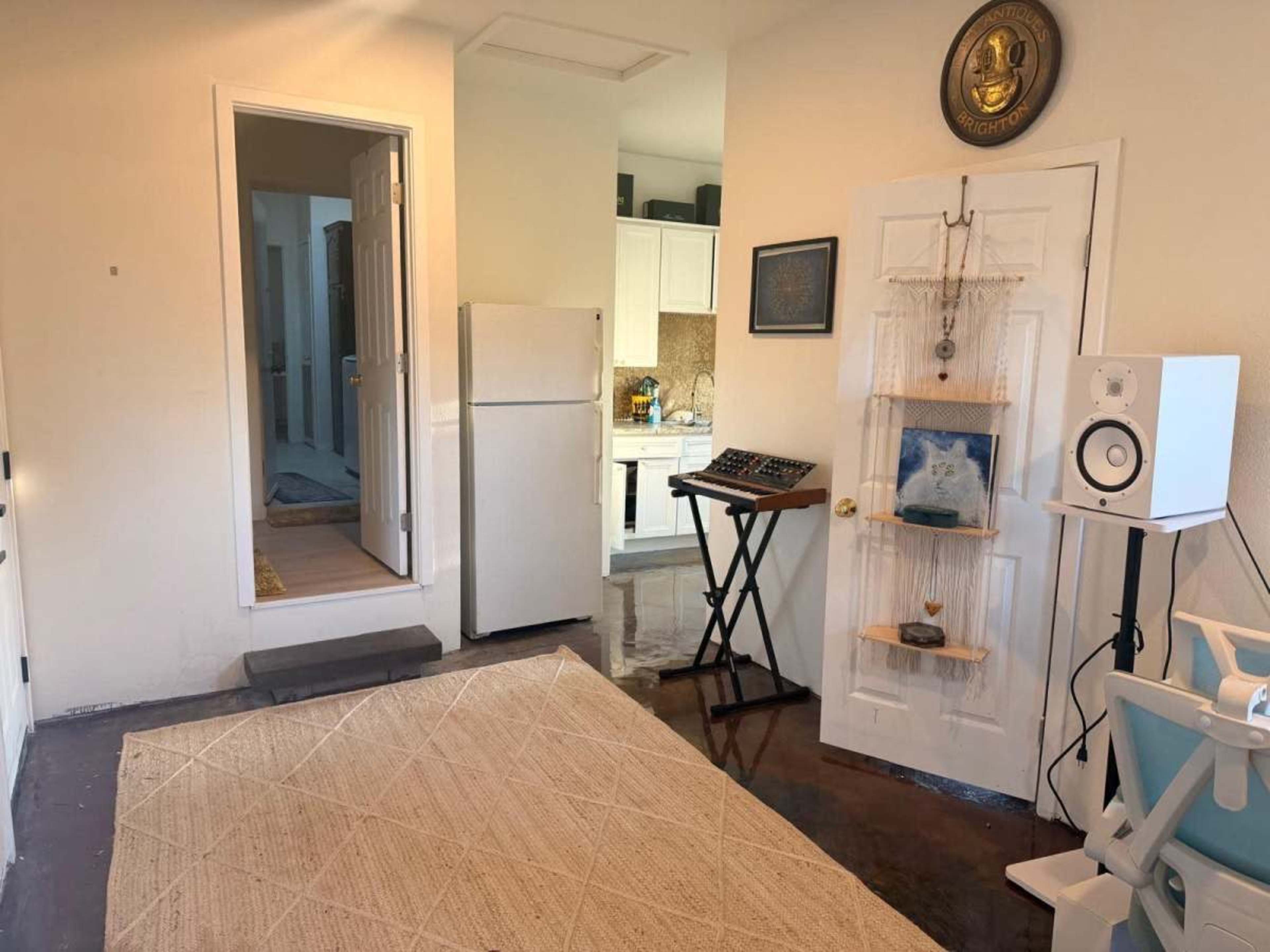 The image shows a small room with a kitchen area visible through an open door, featuring a refrigerator, a keyboard on a stand, and a decorative wall piece near a speaker.