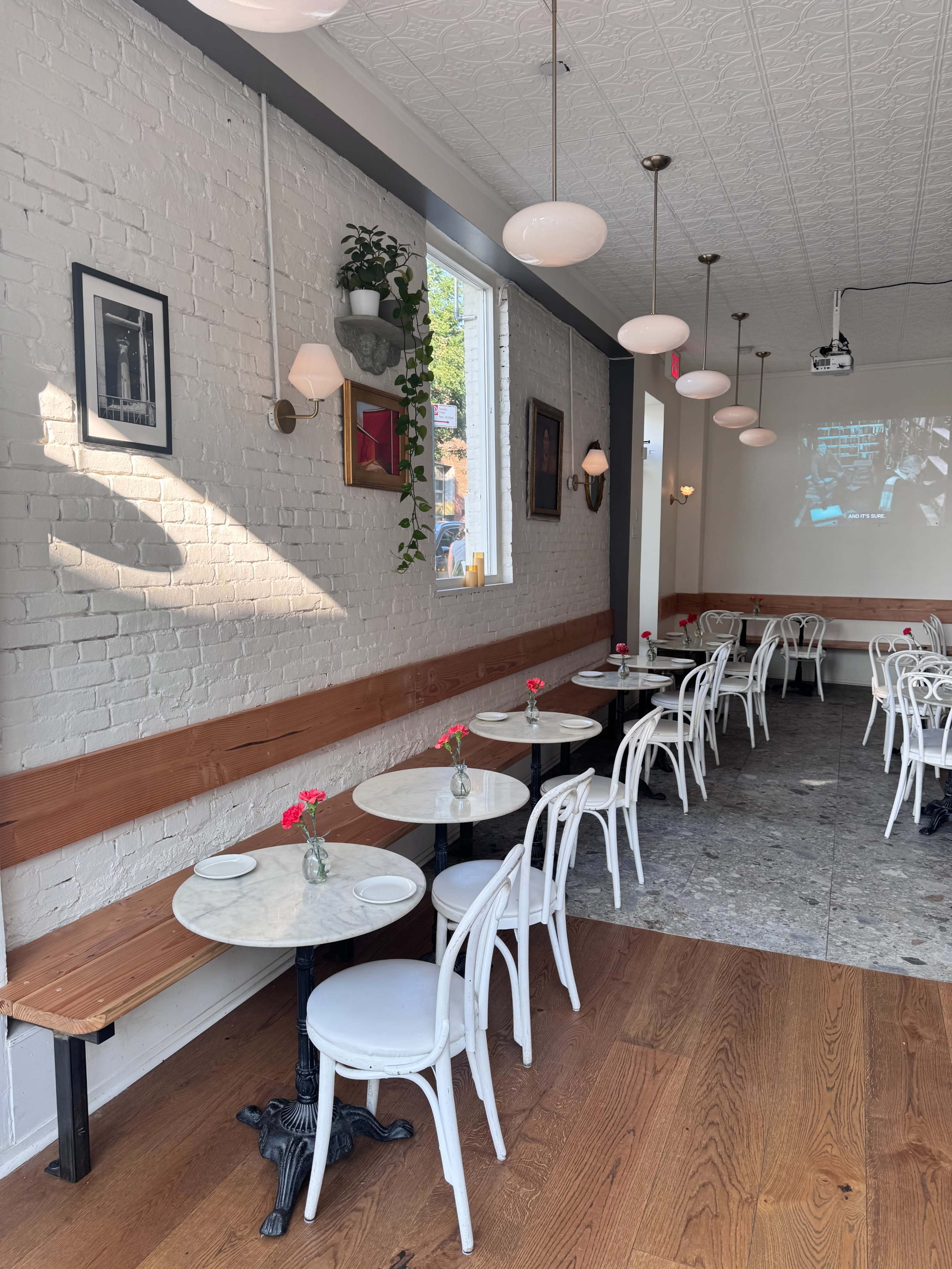 A bright, airy café features white walls, marble-topped tables, and wooden benches alongside a row of white chairs, with small flower arrangements decorating each table.