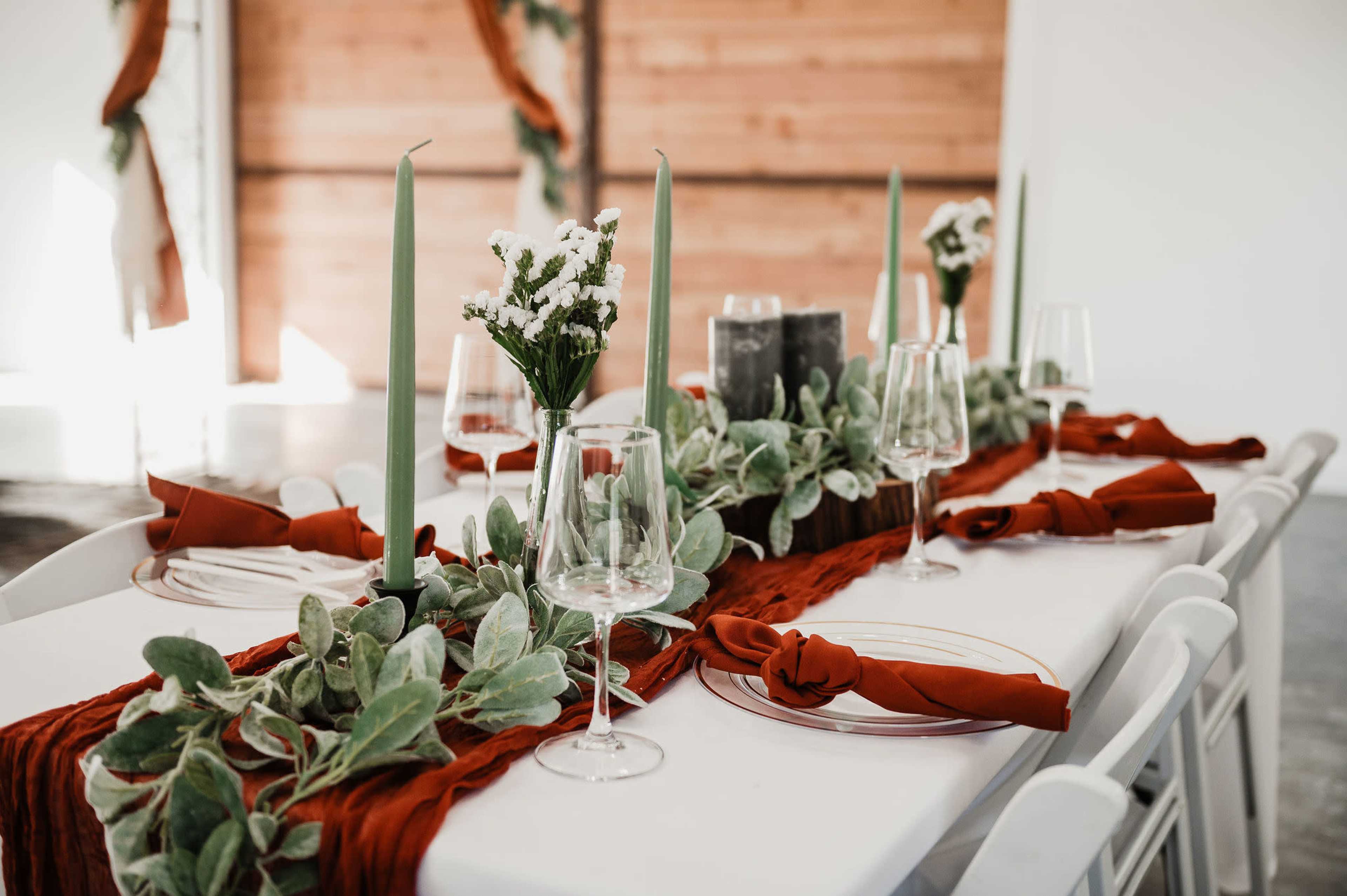 A dining table is set with green candles, floral arrangements, and a red cloth runner, accompanied by glassware and plates.
