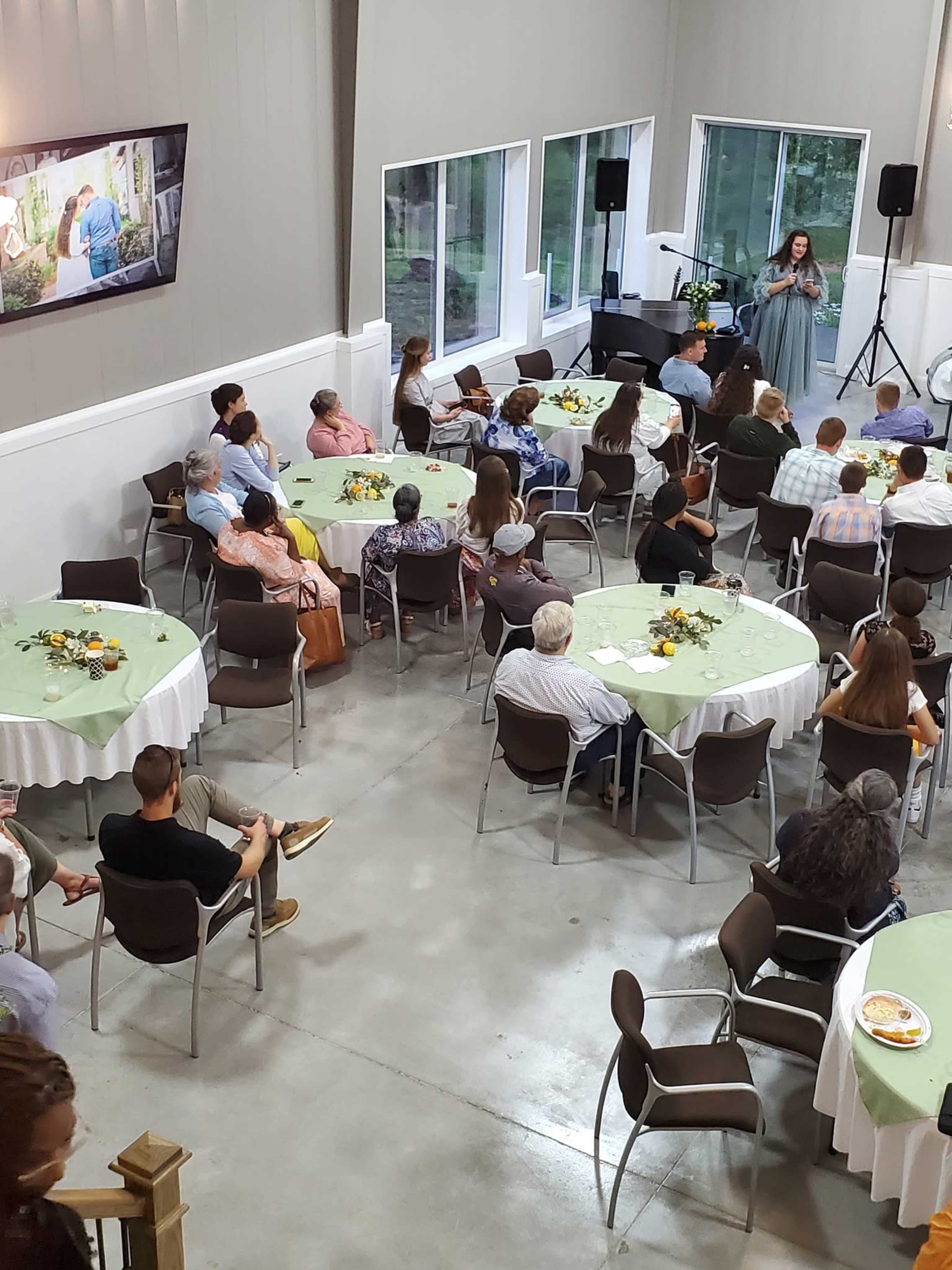 A woman gives a speech in a well-lit reception hall filled with seated guests at round tables.