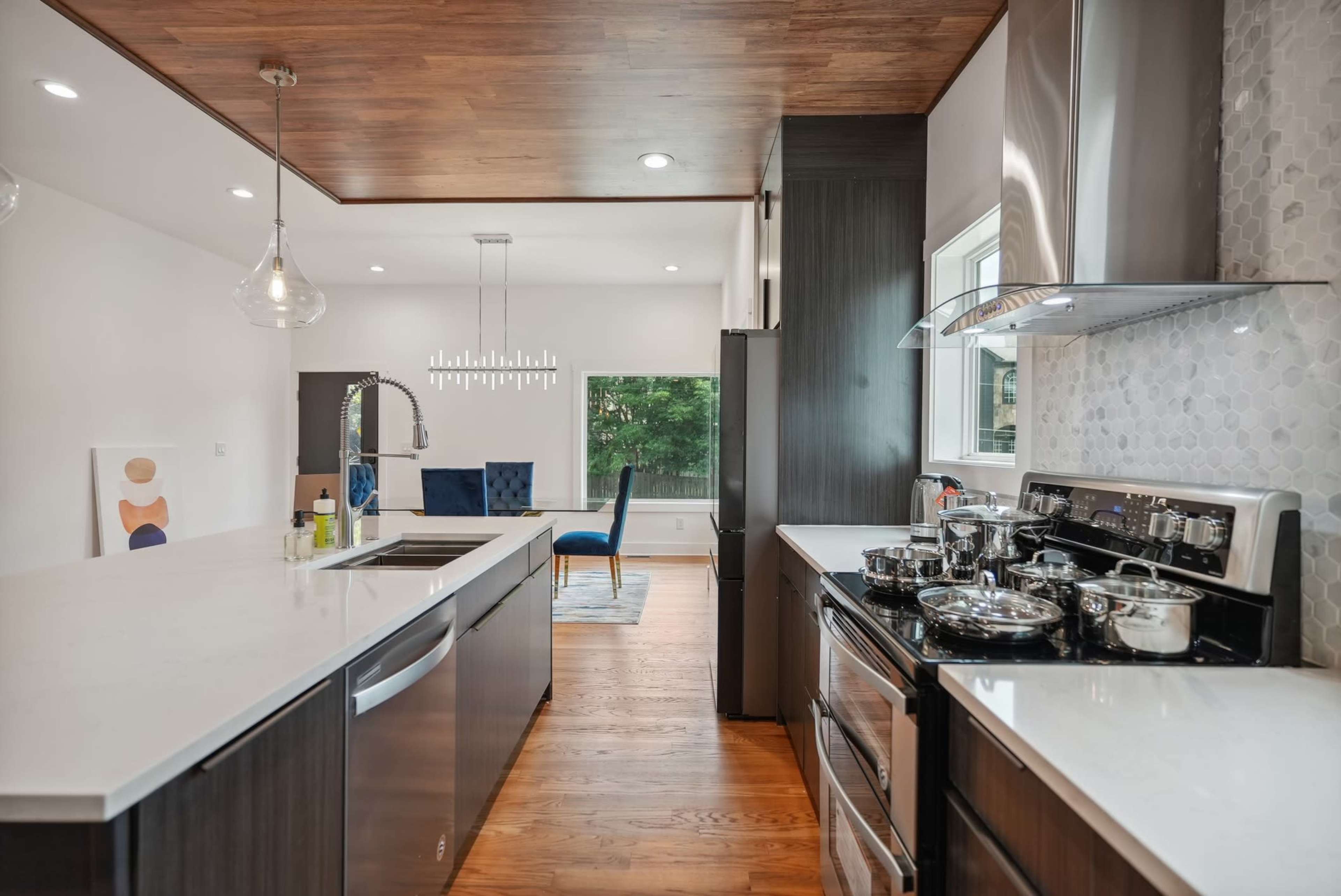 A modern kitchen with dark cabinetry, a white countertop, and stainless steel appliances, leading into a dining area with a blue chair and artwork visible in the background.