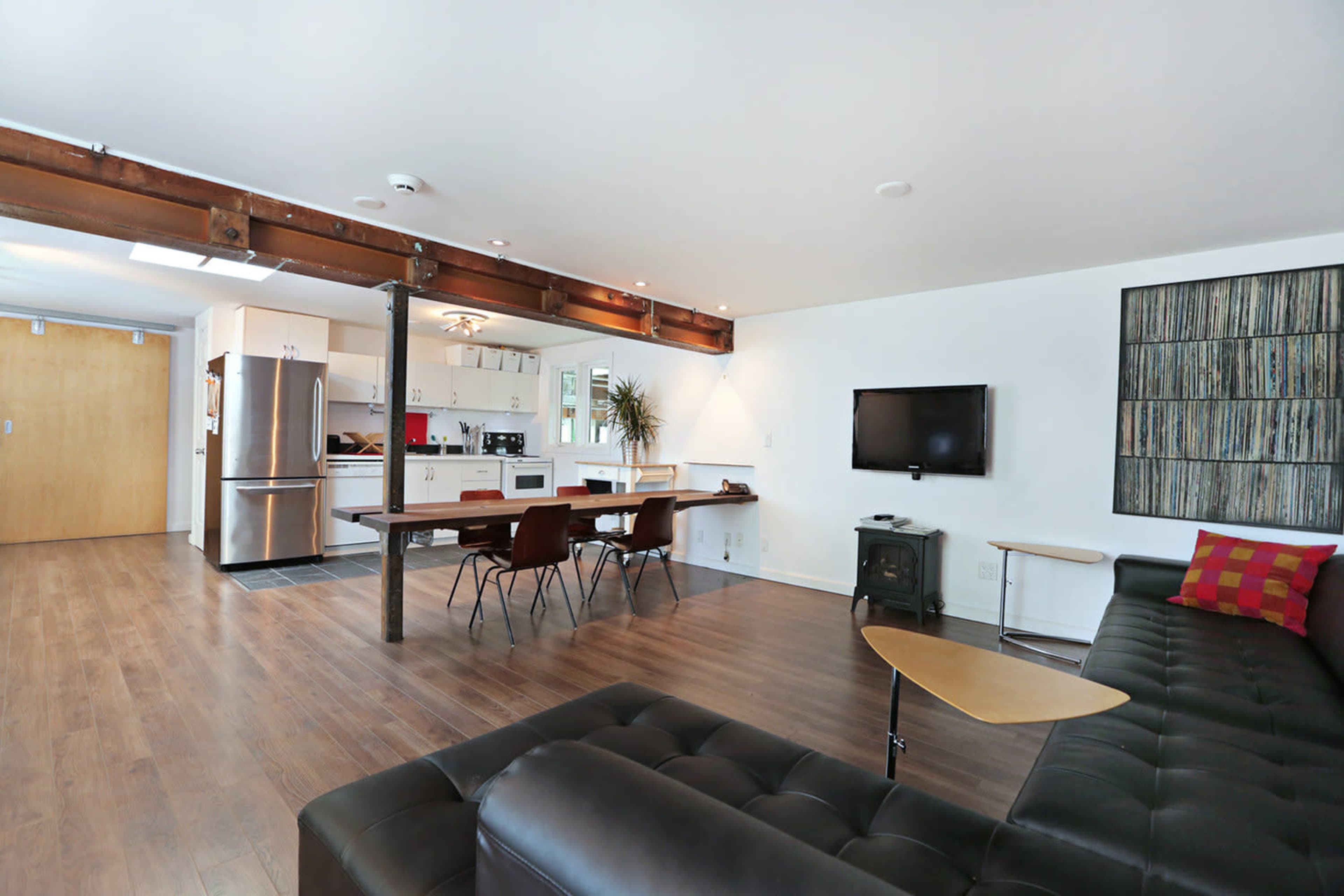 The image shows a modern open-concept living area featuring a kitchen, dining table, and seating arrangement with a wooden floor and minimal decorations.