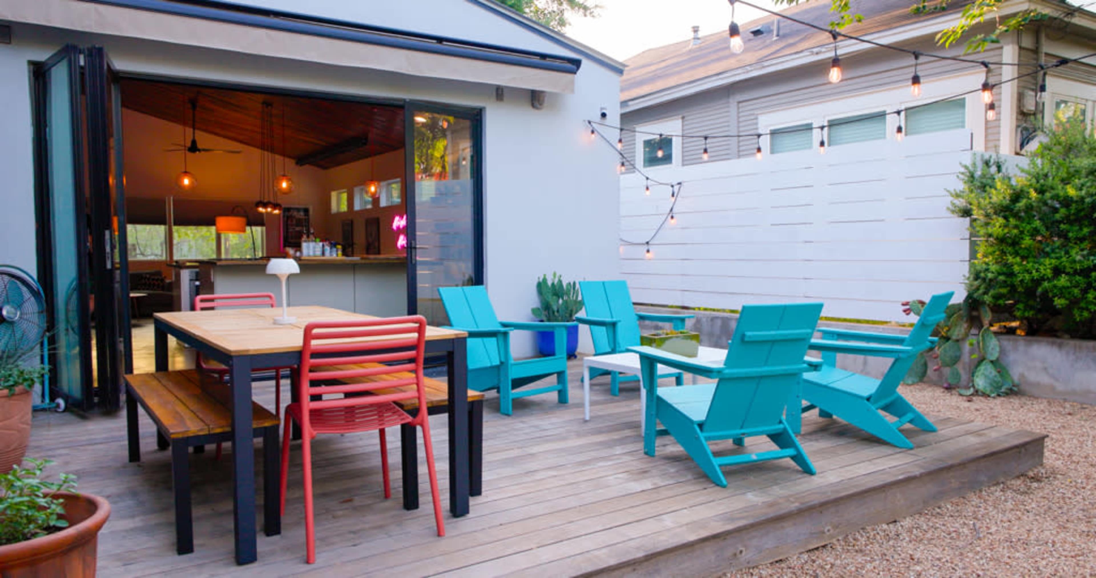 A wooden deck features colorful chairs and a table, surrounded by potted plants and illuminated by string lights.