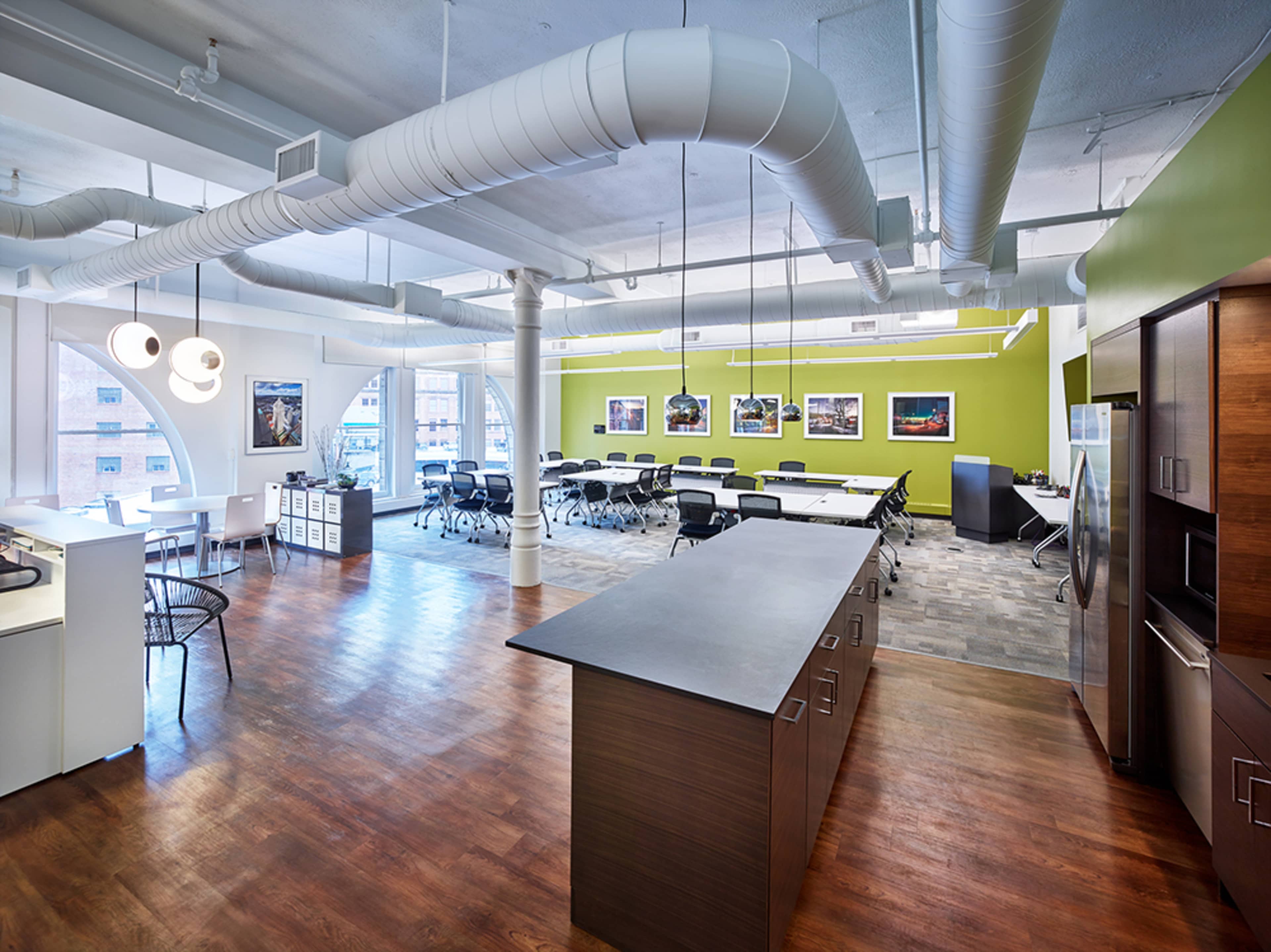 A modern office space featuring a kitchen area with an island, a large meeting area with chairs and tables, and large windows allowing natural light.