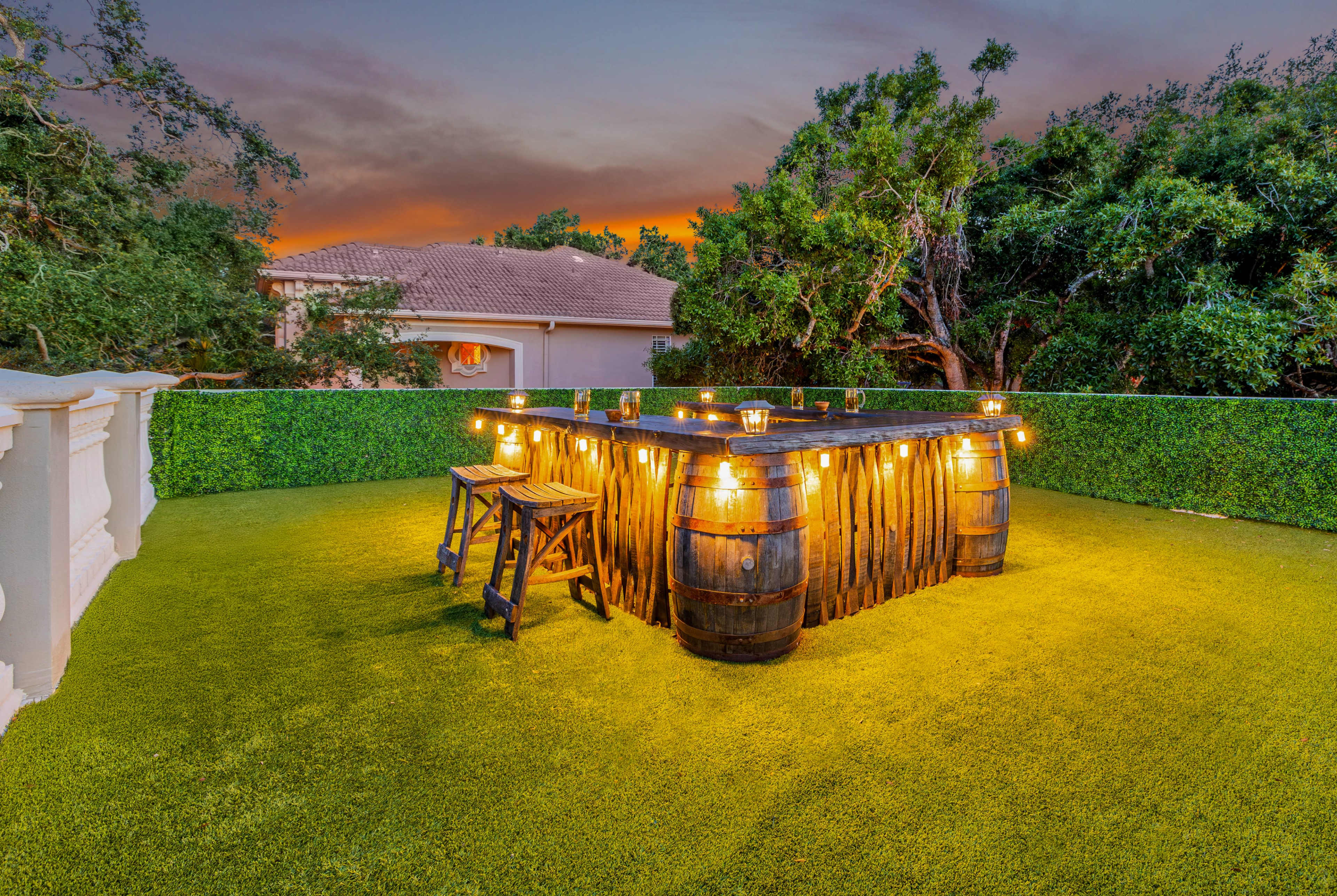 A wooden bar with barrel bases and string lights is situated on a grassy area surrounded by greenery, with a house and sunset in the background.