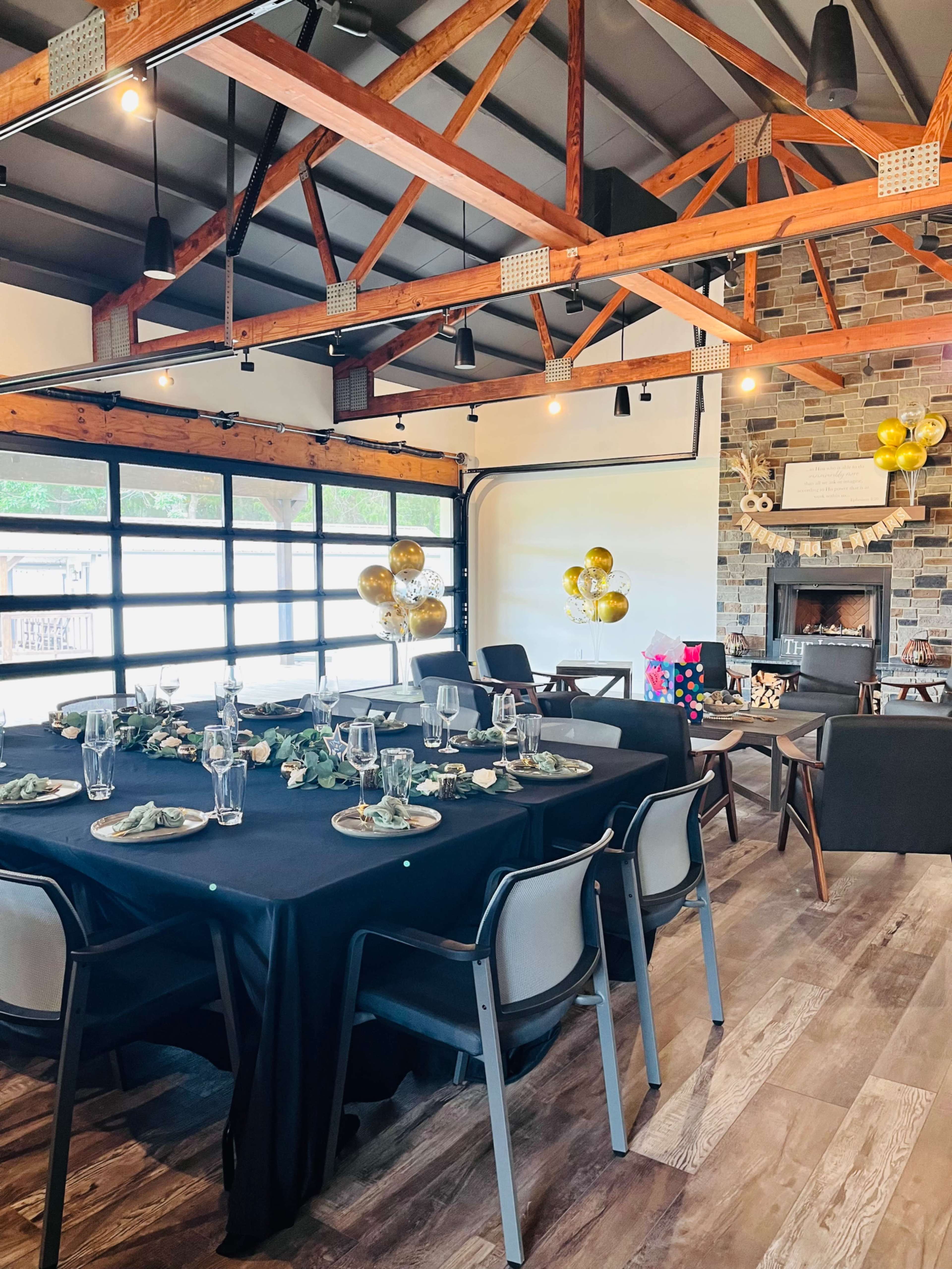 The image shows a decorated dining area with a large black table set with plates and glassware, surrounded by chairs, and a backdrop of a stone fireplace and balloons.