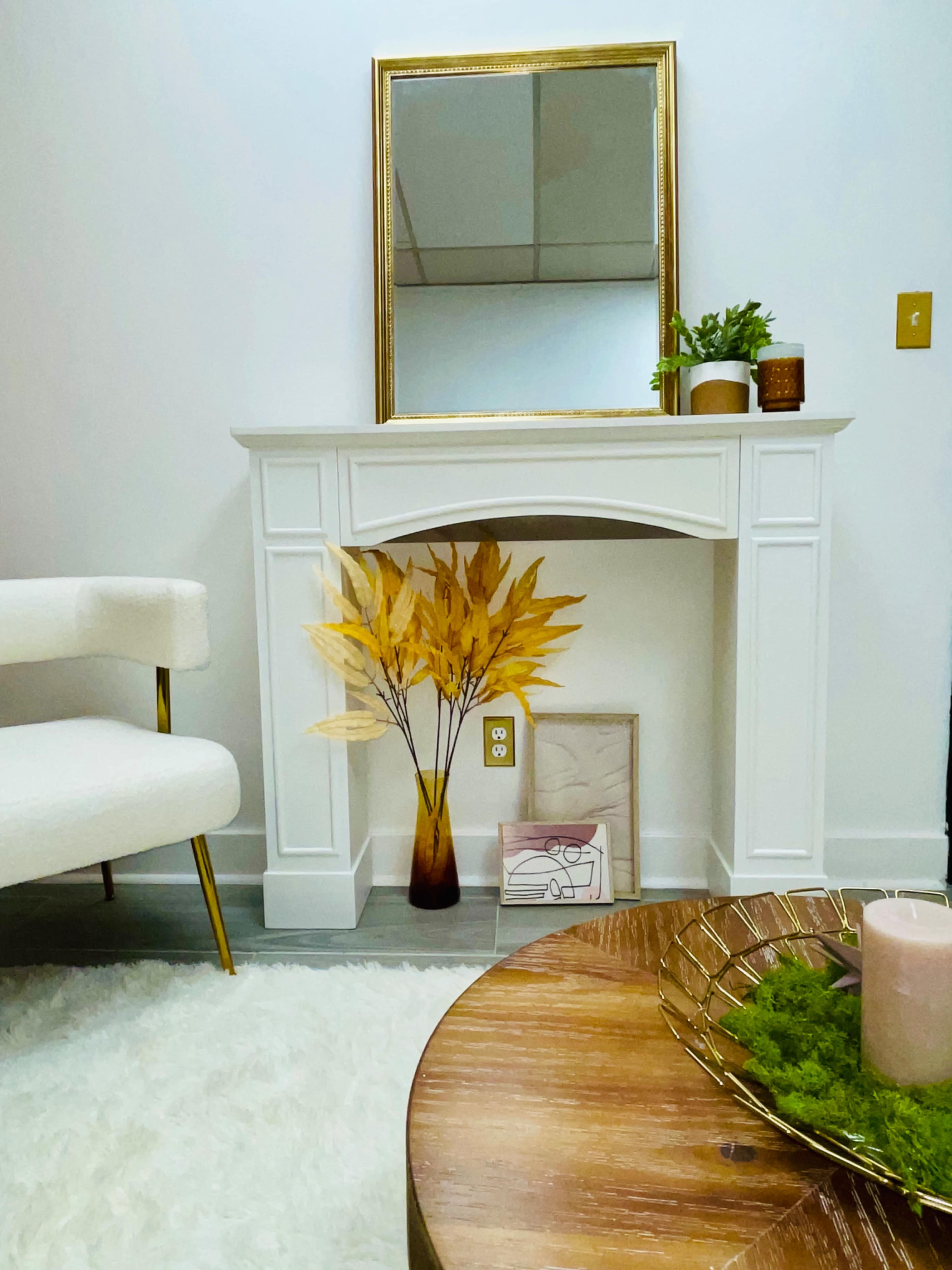 A white mantel with a mirror above it is decorated with a vase of dried leaves, a small plant, and a few framed artworks, while a wooden coffee table and a light-colored chair are visible in the foreground.