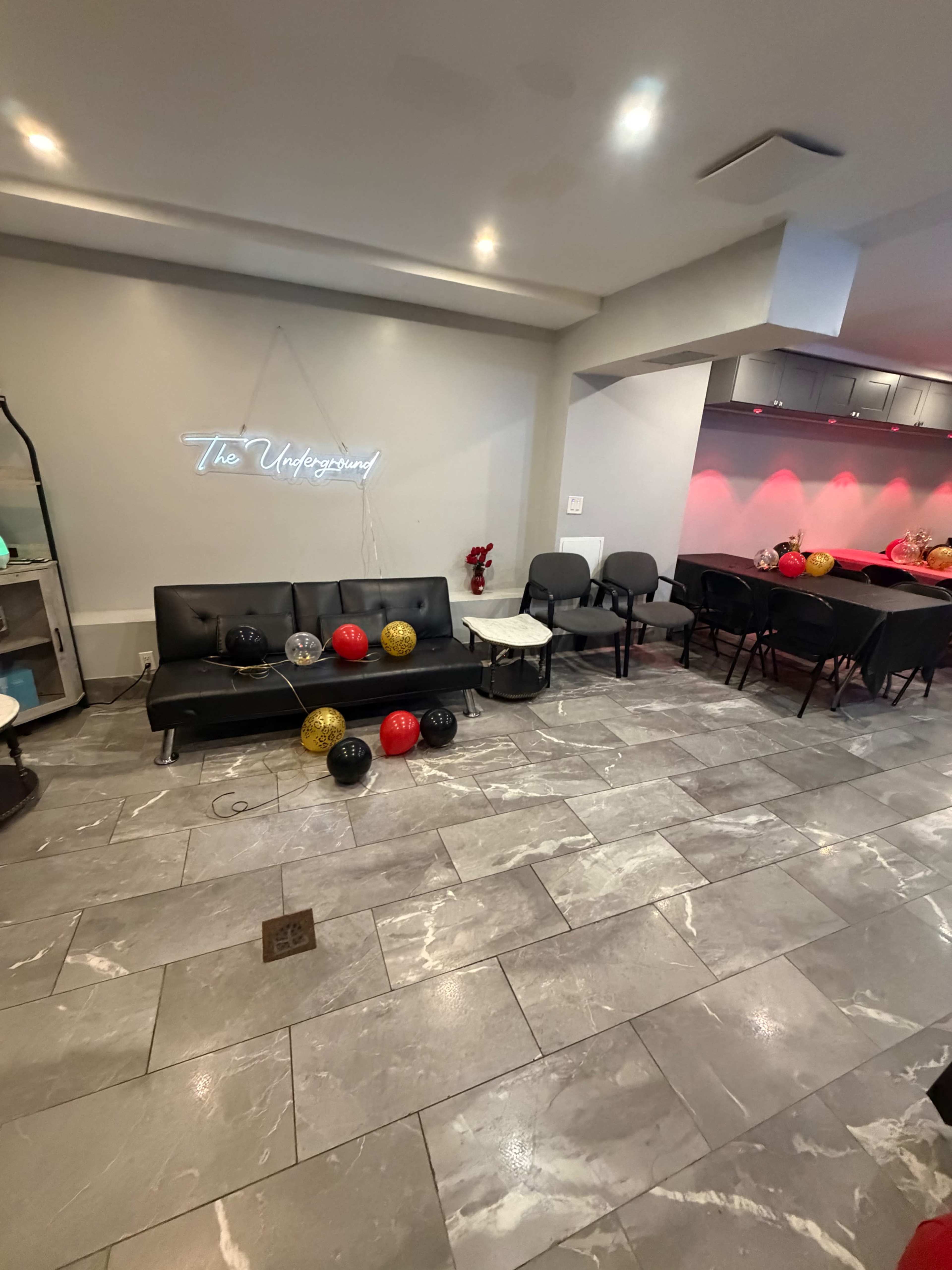 The image shows a modern lounge area with black couches, a small table, and colorful balloons scattered on the gray tiled floor.