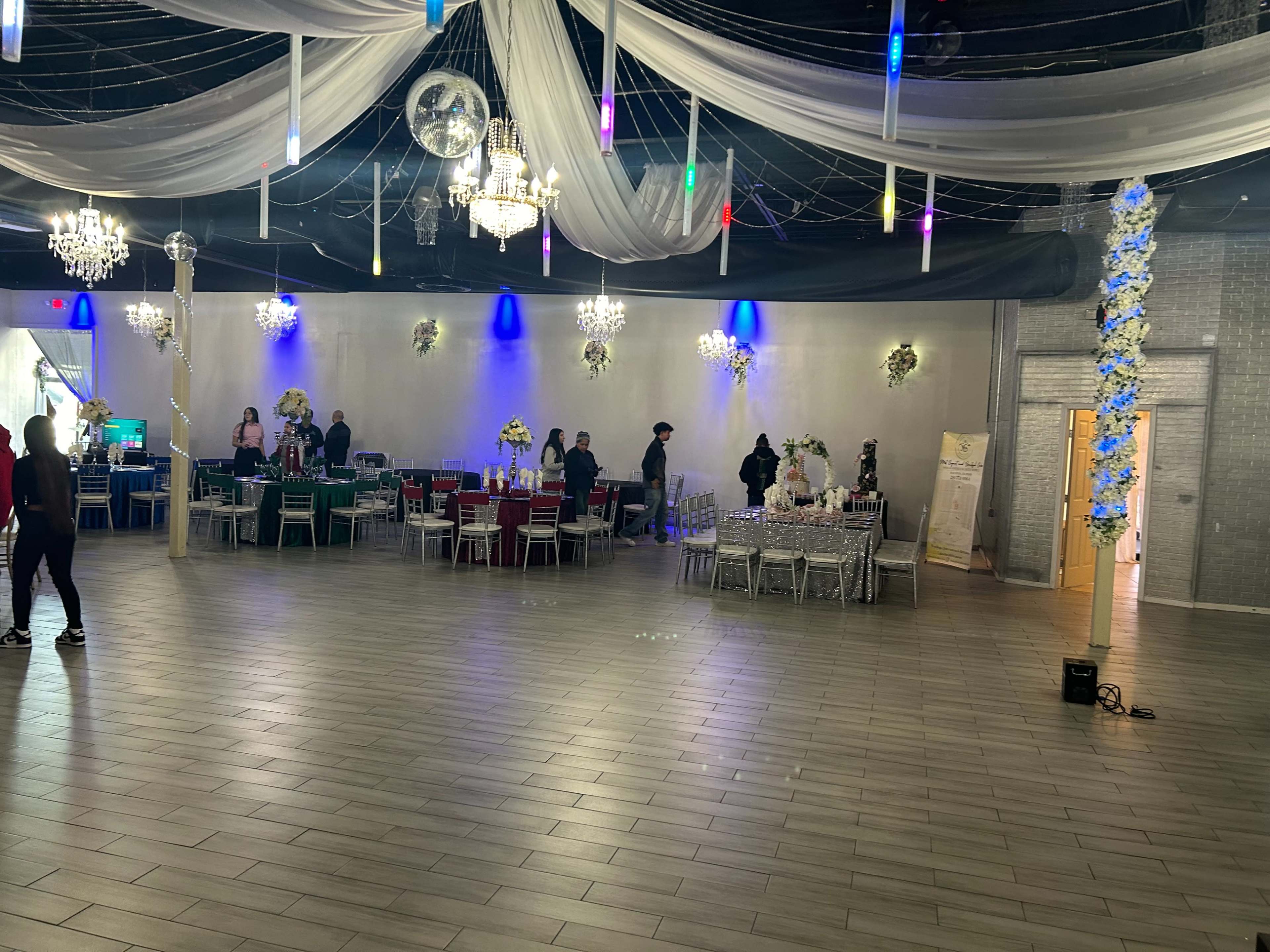 A spacious event venue with decorated tables set up for a gathering, featuring colorful lighting and chandeliers.