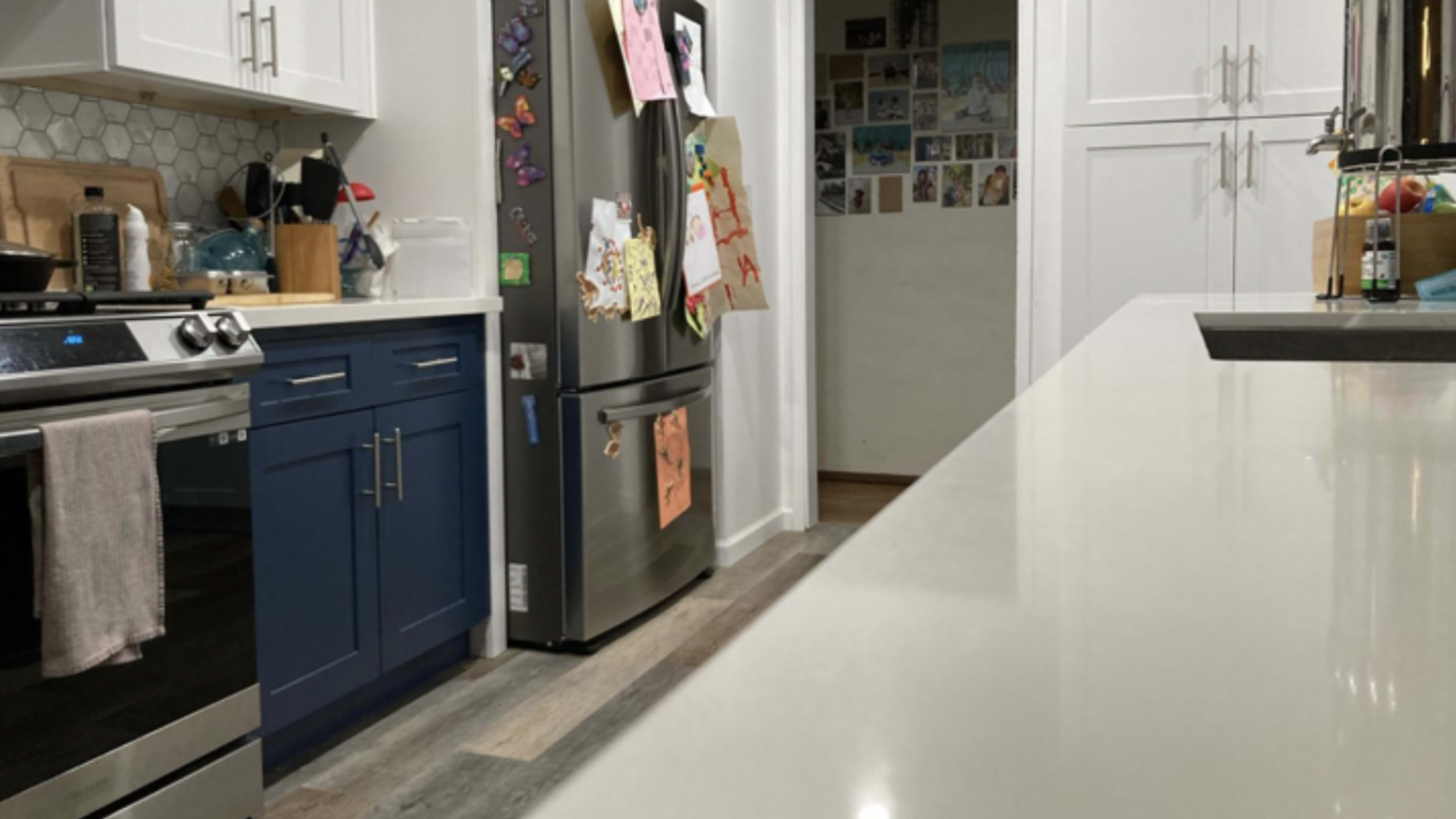 A kitchen with blue cabinetry, a stainless steel refrigerator, and a countertop leading to a hallway adorned with various images.