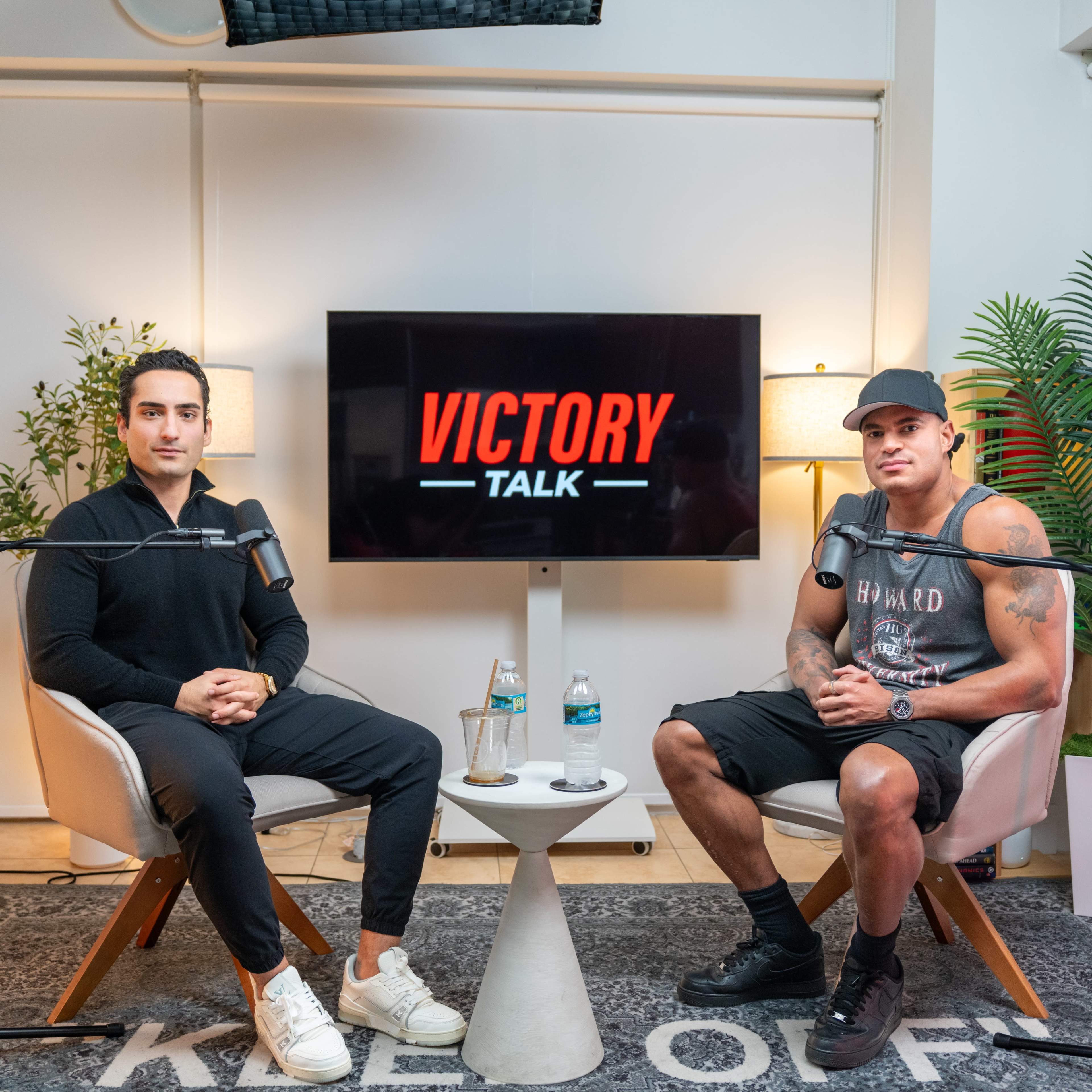 Two men sit in a podcast studio, each in a chair with microphones, facing the camera, with a screen displaying "VICTORY TALK" in the background.