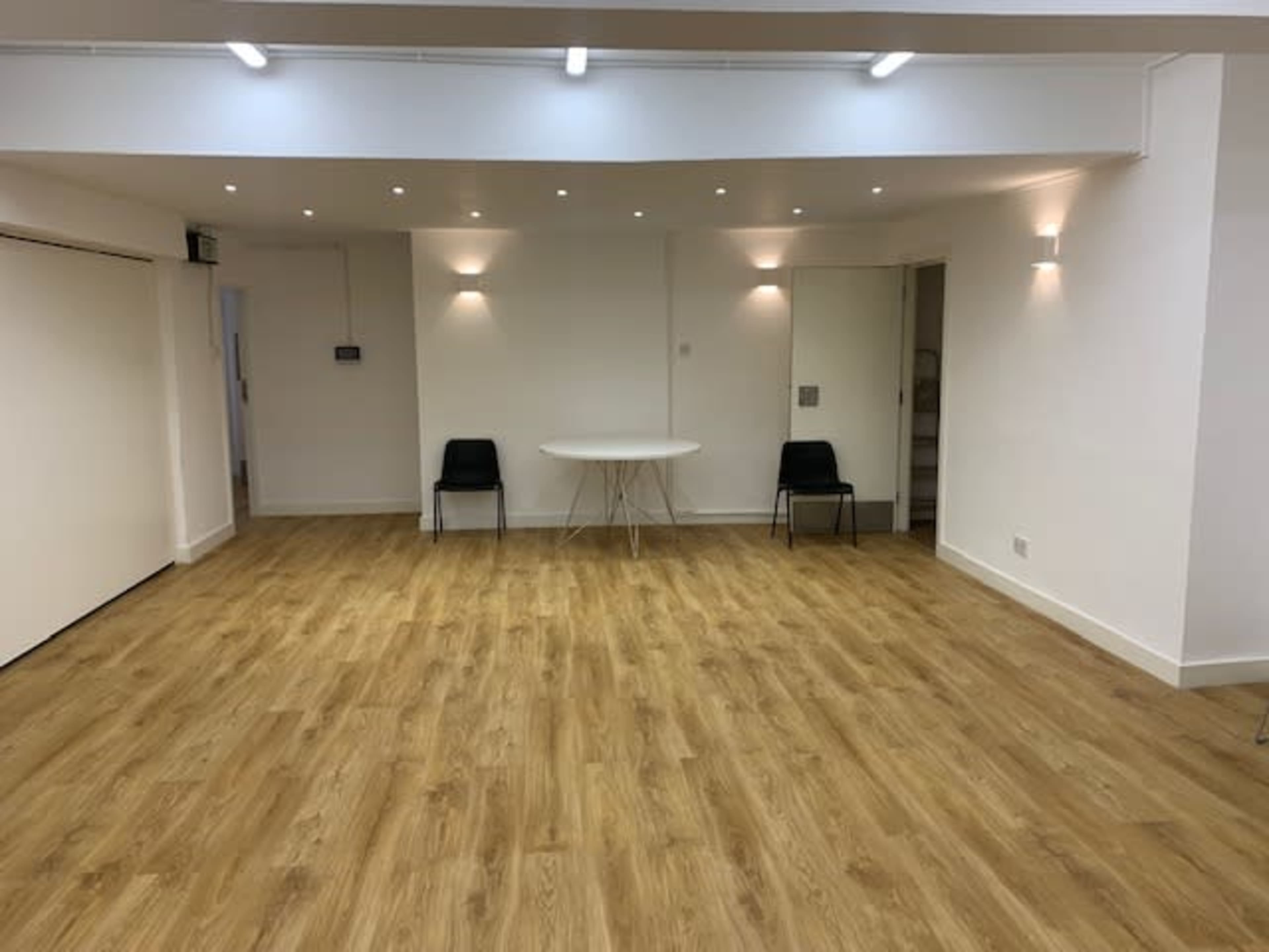 A spacious room with a light wooden floor, a round table in the center, and two black chairs placed on opposite sides.