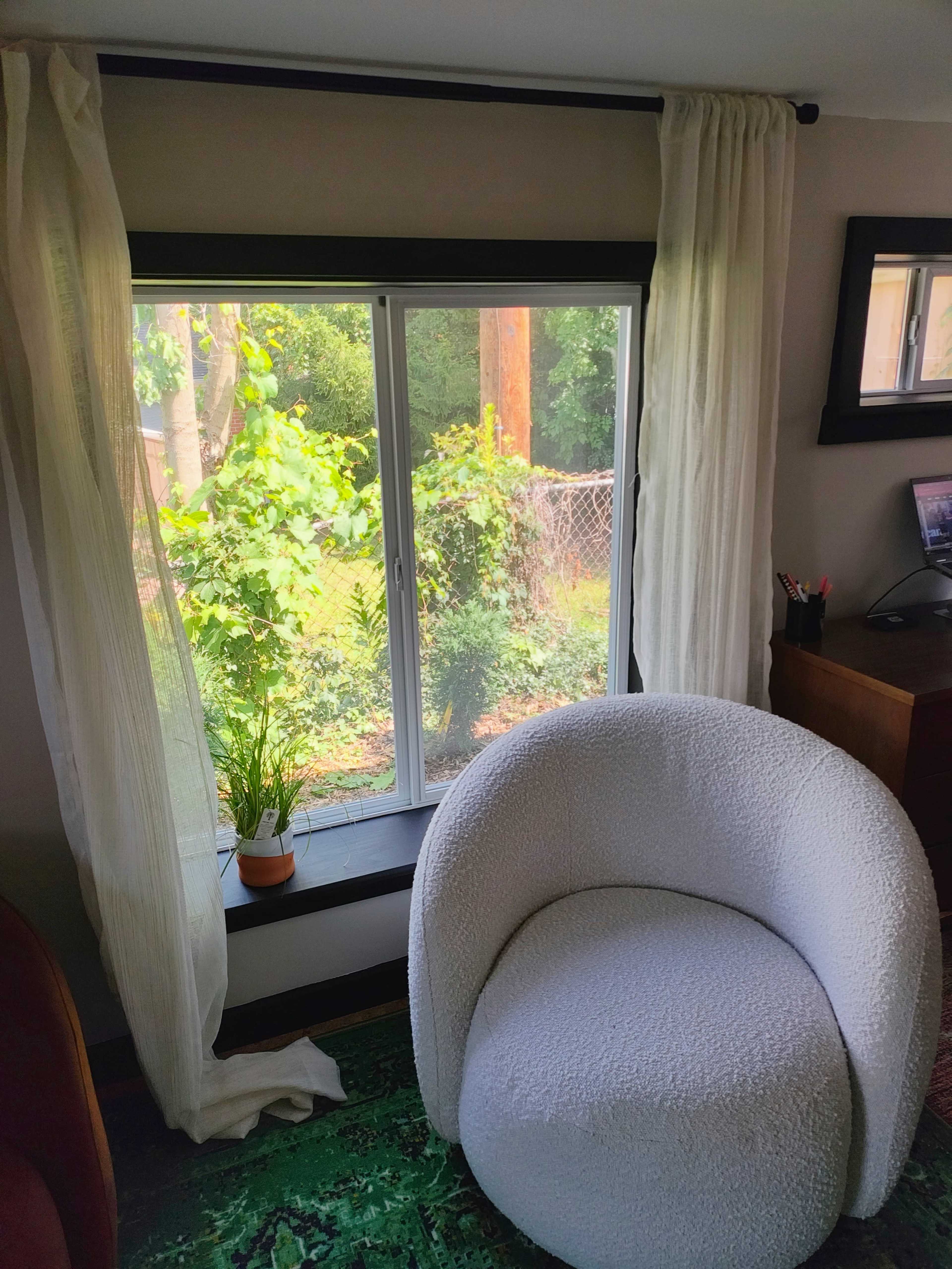 A circular, white upholstered chair sits in front of a large window adorned with sheer curtains, overlooking a green outdoor space.