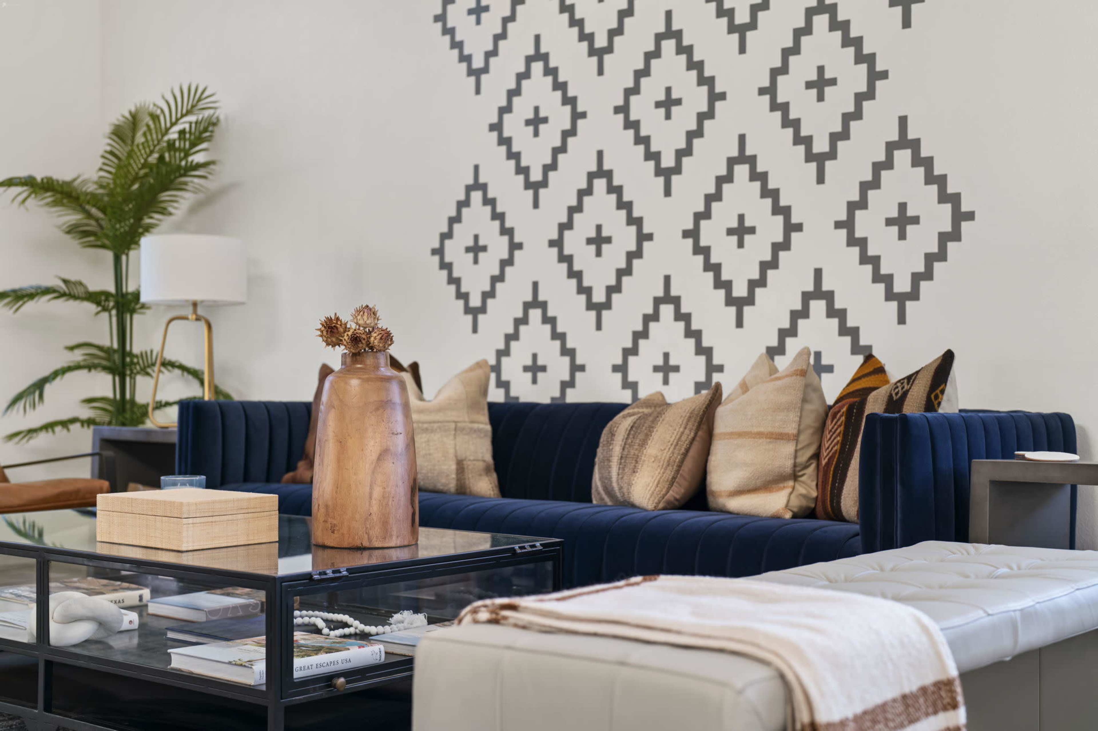 A modern living room features a navy blue velvet sofa adorned with patterned pillows, a glass coffee table, and a decorative vase on a side table against a geometric wall design.