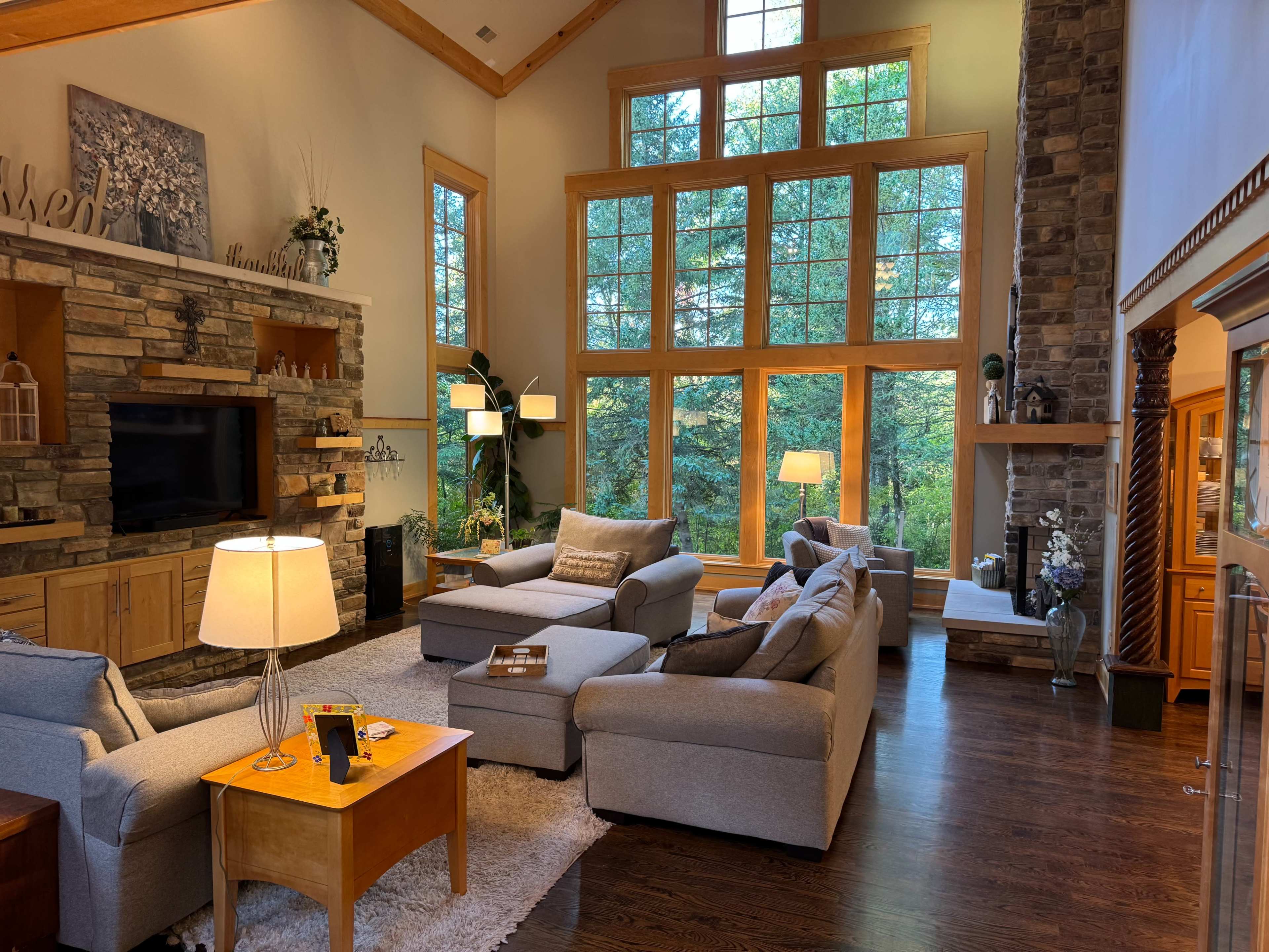 The image shows a spacious living room with large windows, a stone fireplace, and comfortable gray furniture arranged around a wooden coffee table.