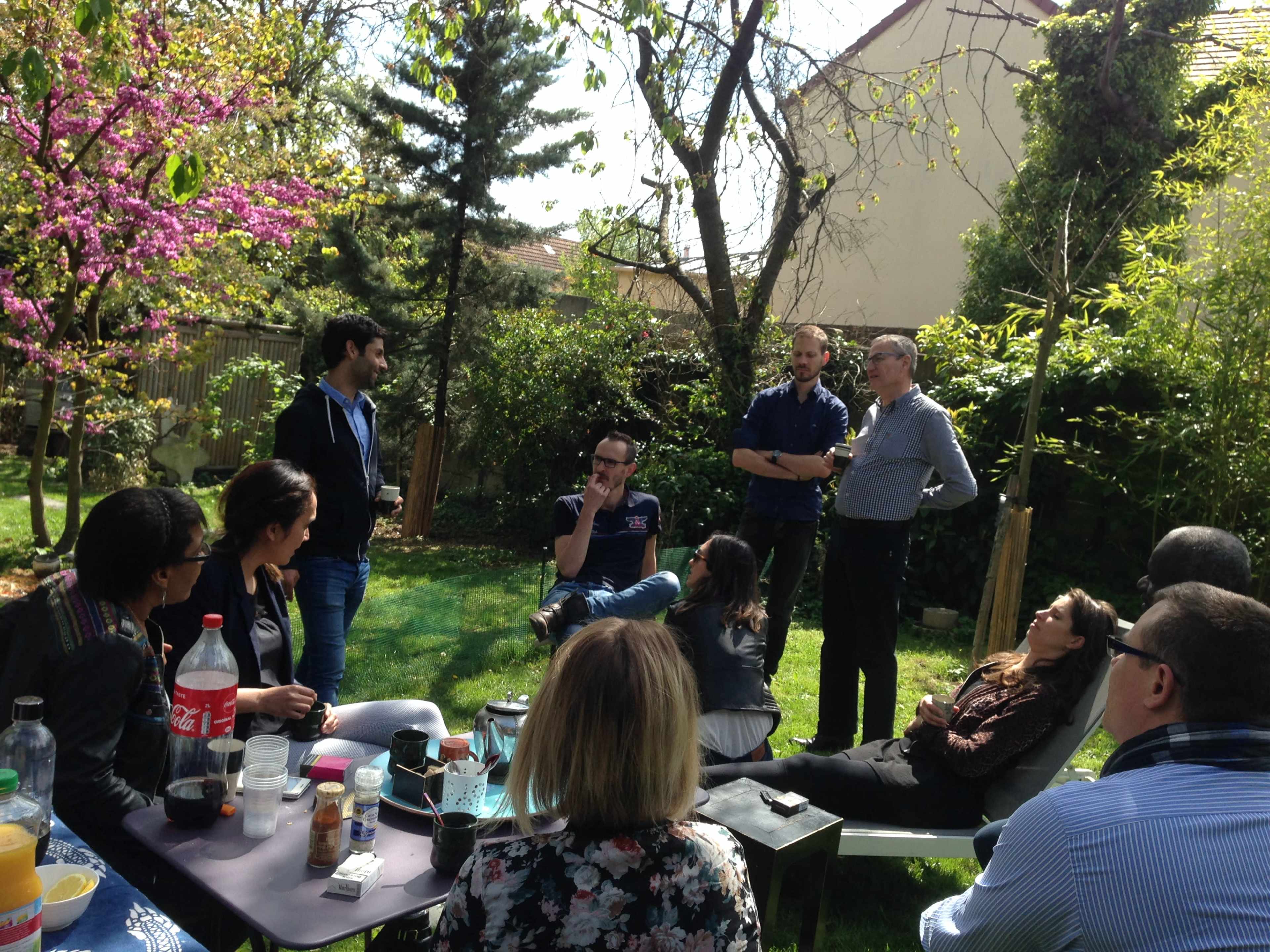A group of people is gathered in a garden, engaging in conversation while seated and standing among greenery and blooming trees.