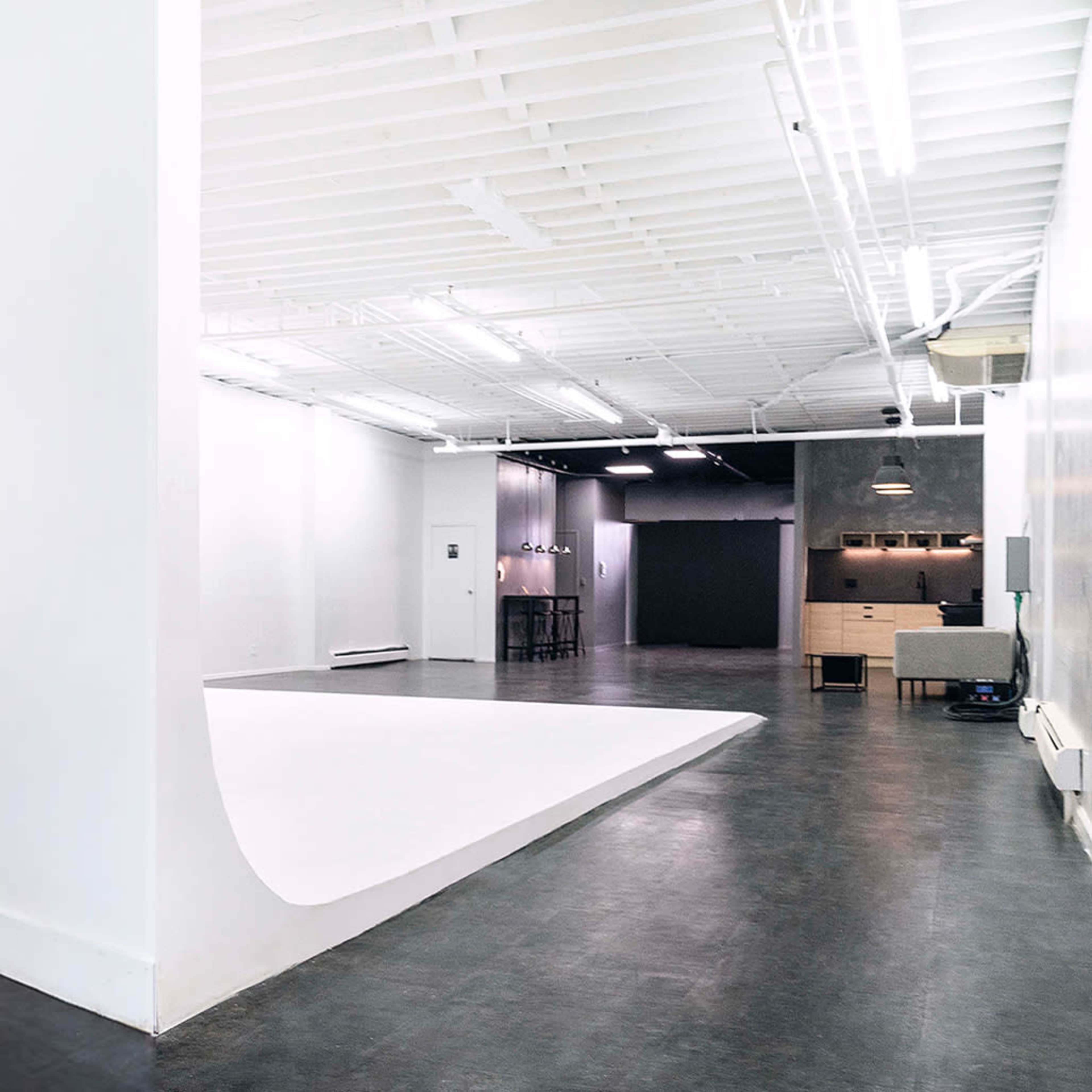 A spacious studio with white walls, a large white backdrop, and a minimalist design featuring a seating area and a small kitchenette.