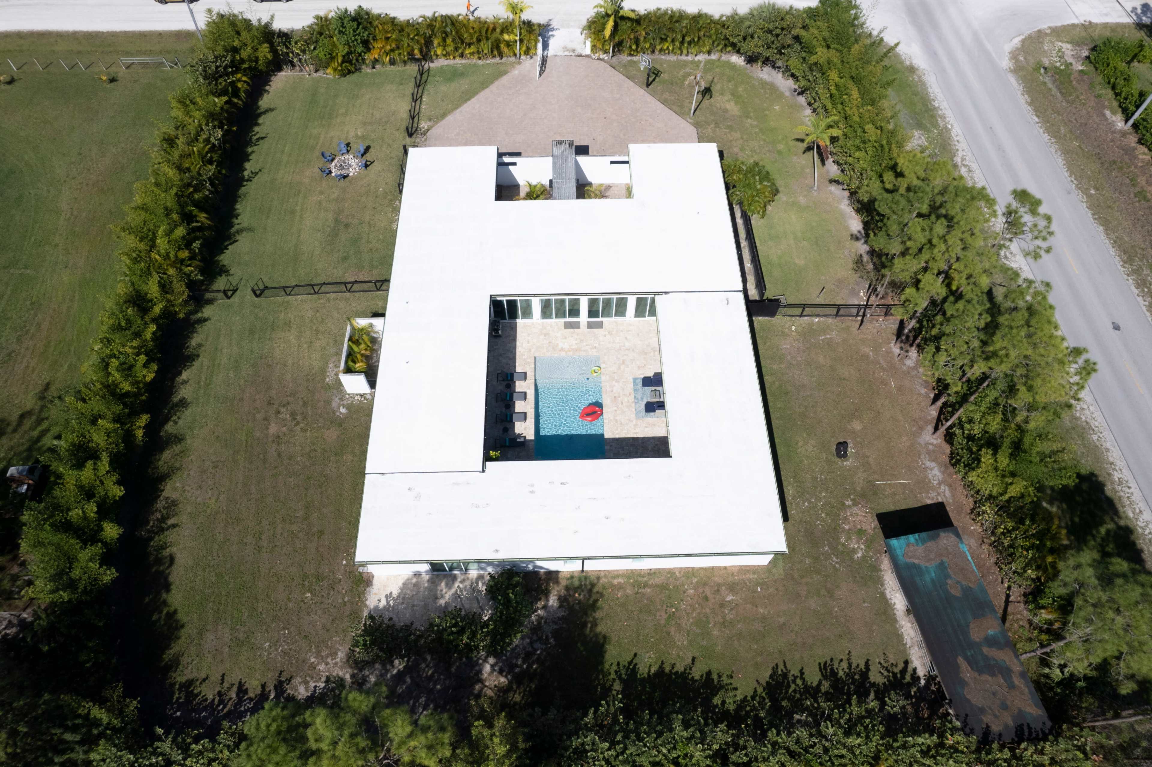 An aerial view shows a modern white building with a central pool surrounded by a gravel patio and bordered by greenery, located near a road.