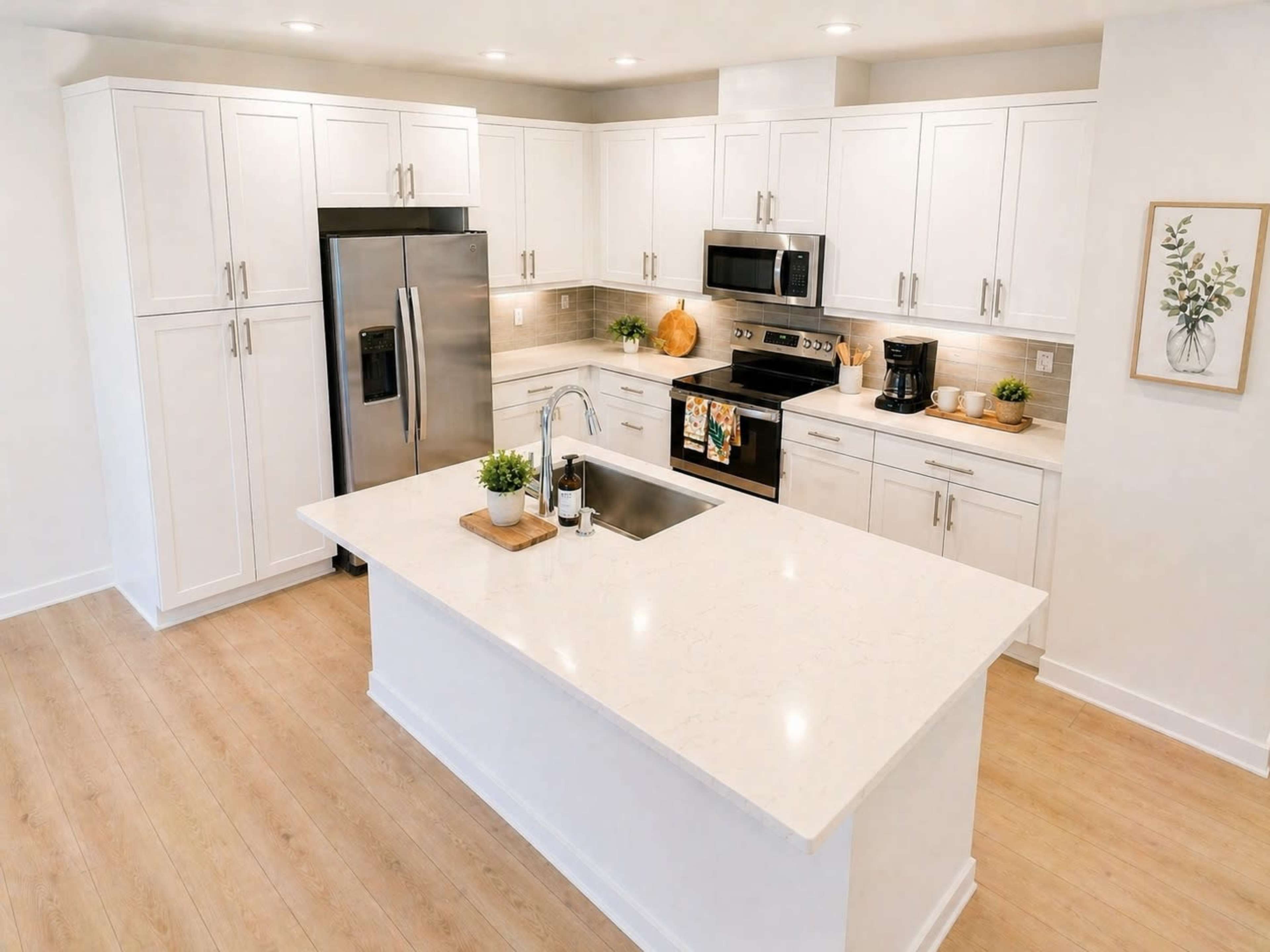 The image shows a modern kitchen featuring white cabinetry, stainless steel appliances, a large island with a sink, and wooden flooring.
