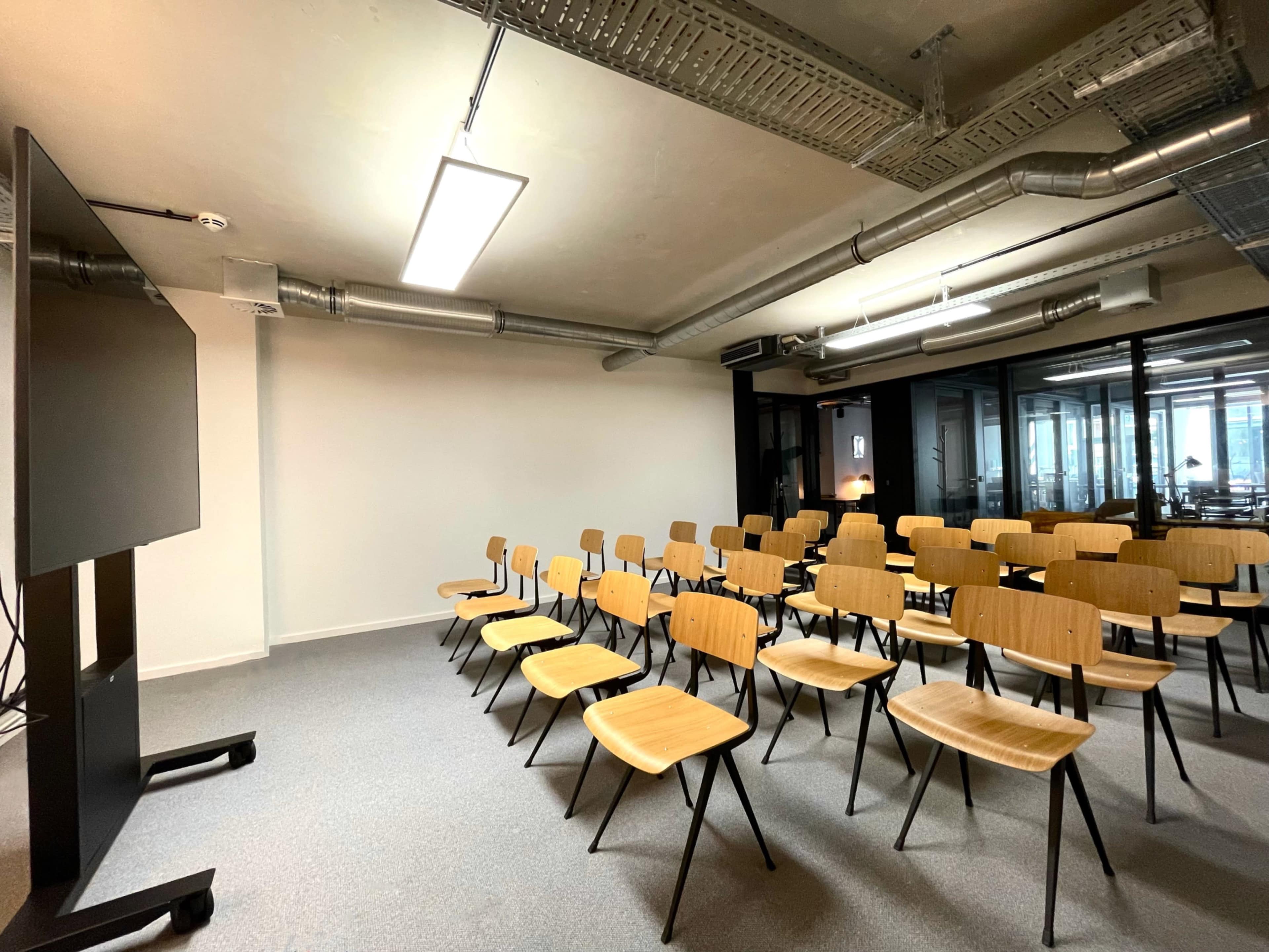 A row of empty wooden chairs faces a large wall-mounted screen in a modern office space.
