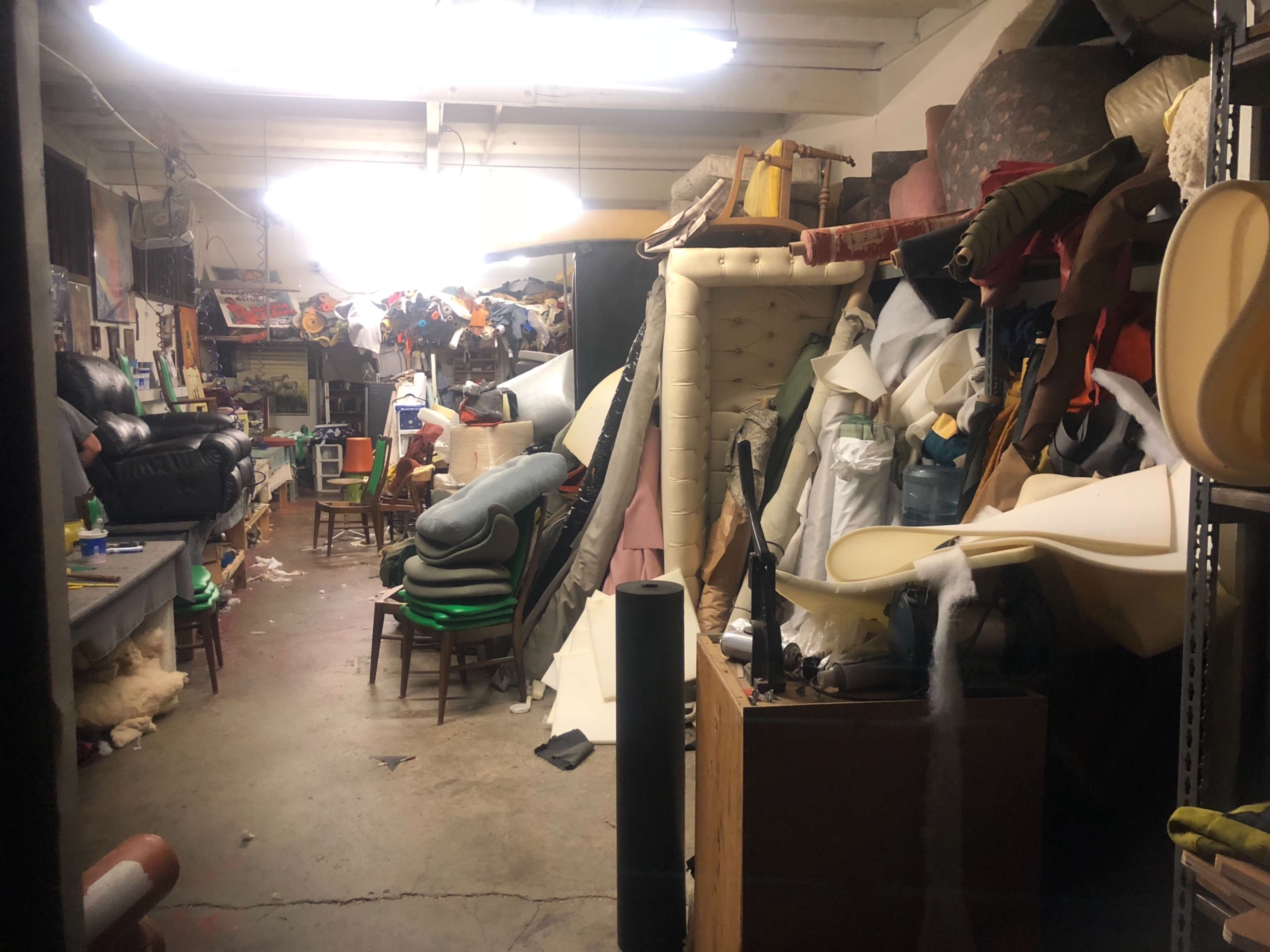 A cluttered workshop filled with various types of upholstery materials, furniture pieces, and tools.