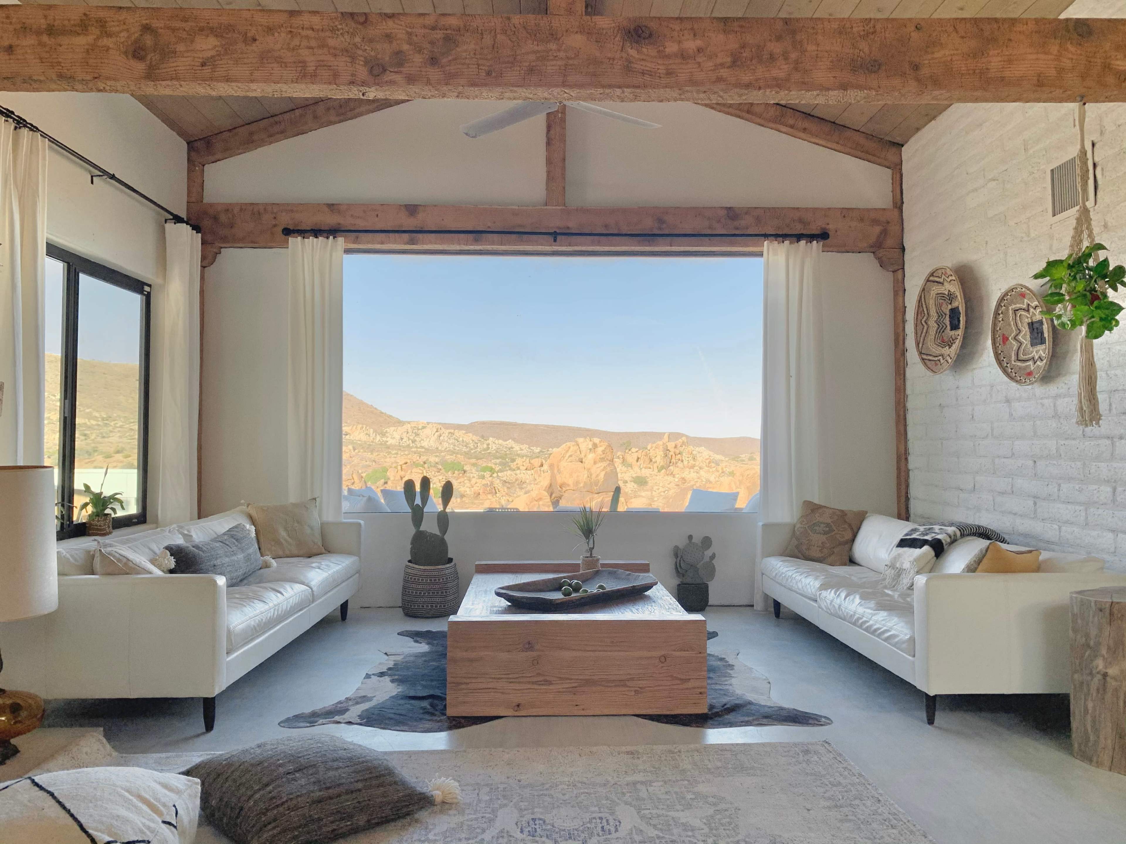 The living room features two white sofas facing a wooden coffee table, with large windows showcasing a mountainous landscape in the background.