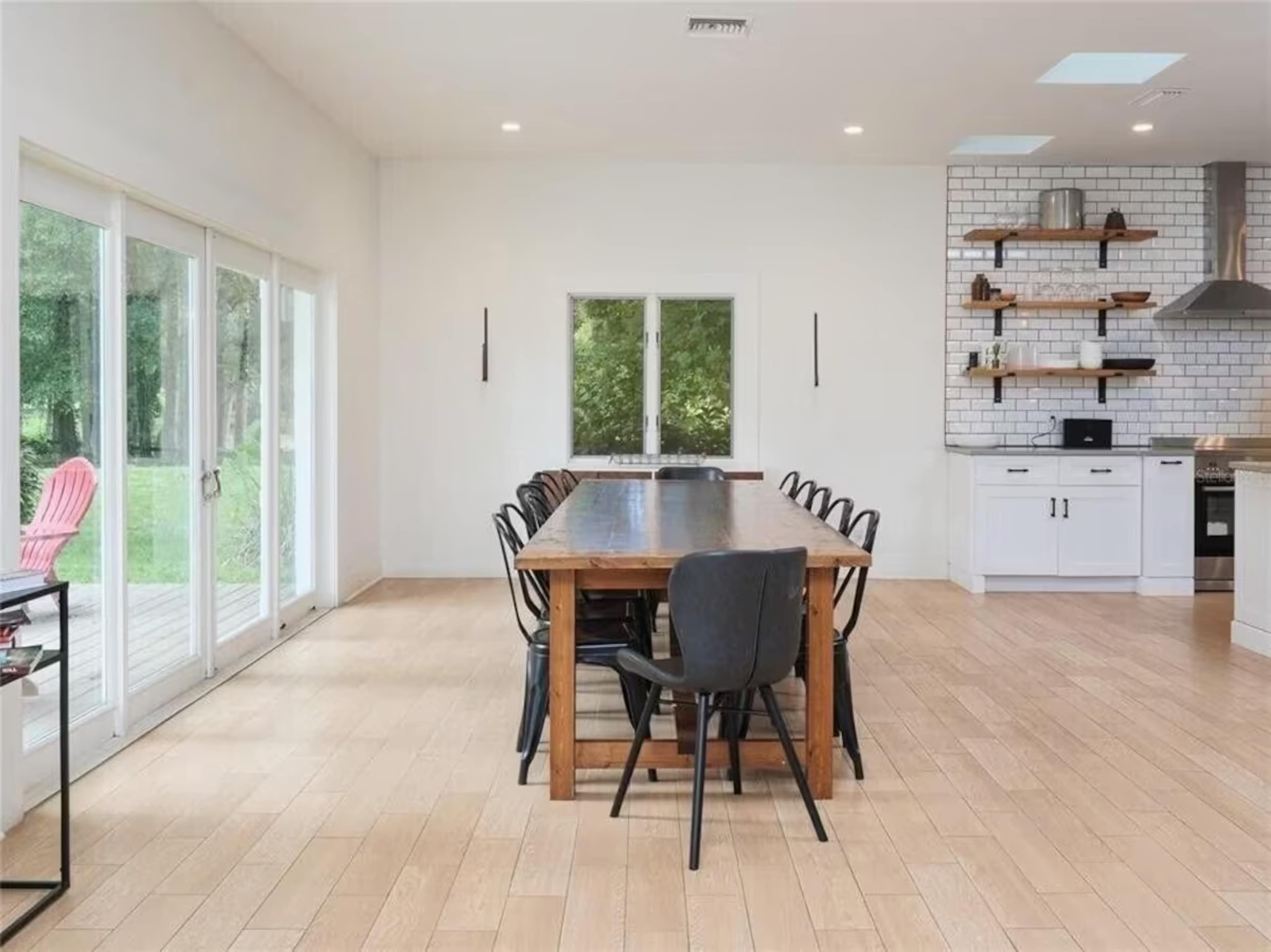 A spacious dining area with a large wooden table surrounded by black chairs and large windows overlooking a green outdoor space.