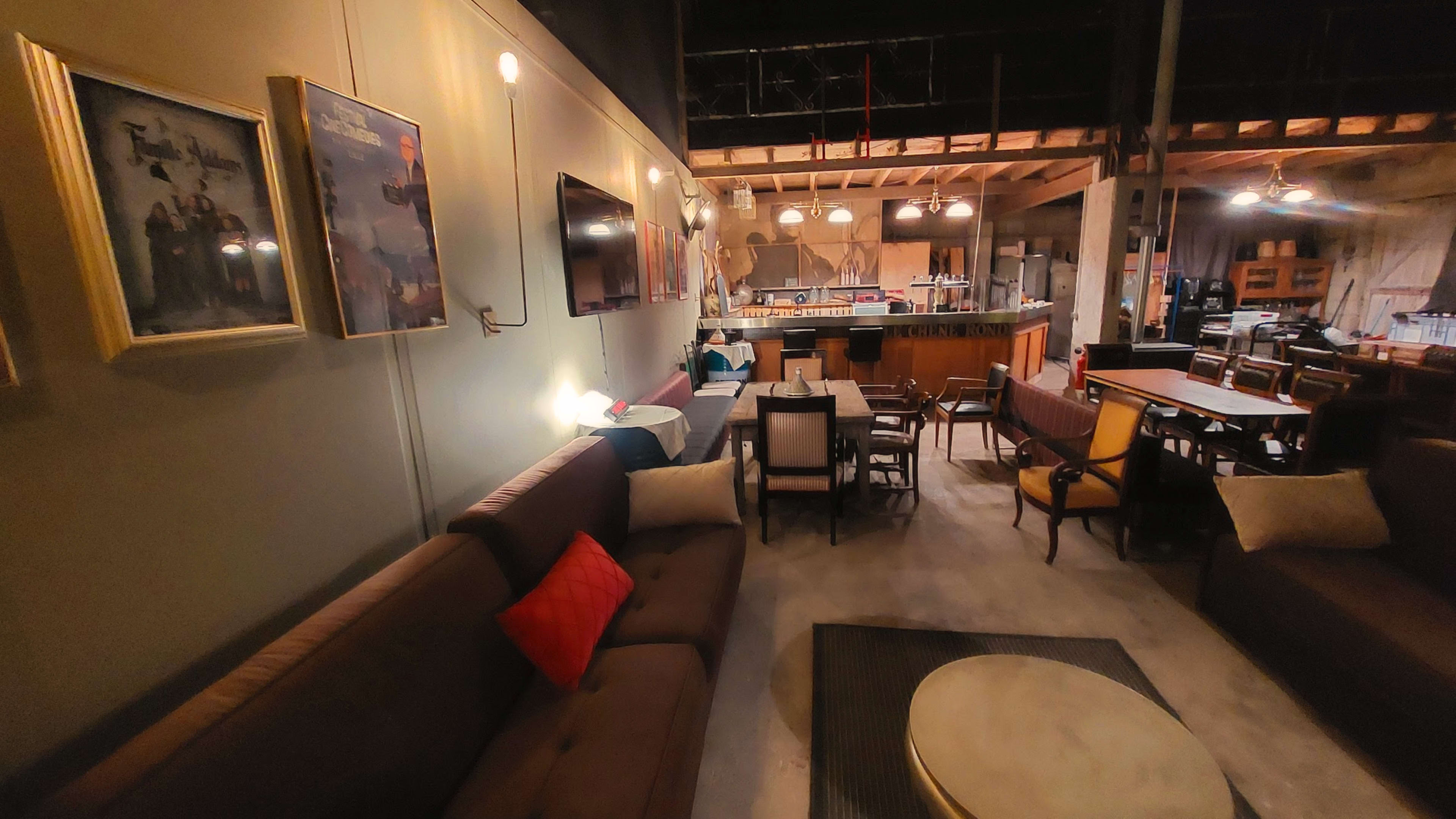 The image shows a spacious, dimly lit lounge area with various seating arrangements, a bar in the background, and framed artwork on the walls.