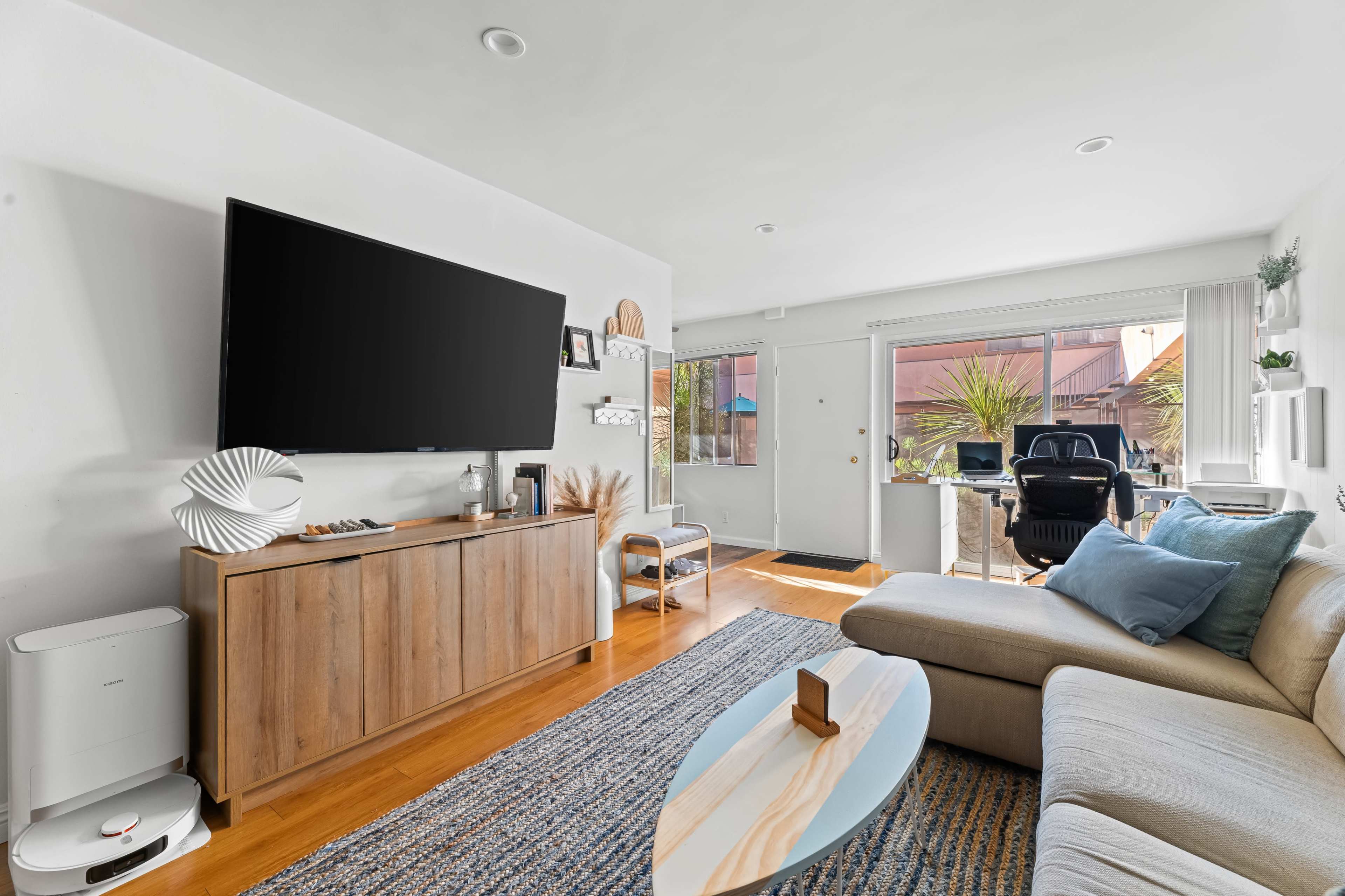The living room features a large television on a wooden cabinet, a light-colored sectional sofa, and a coffee table, with natural light coming in through a window.