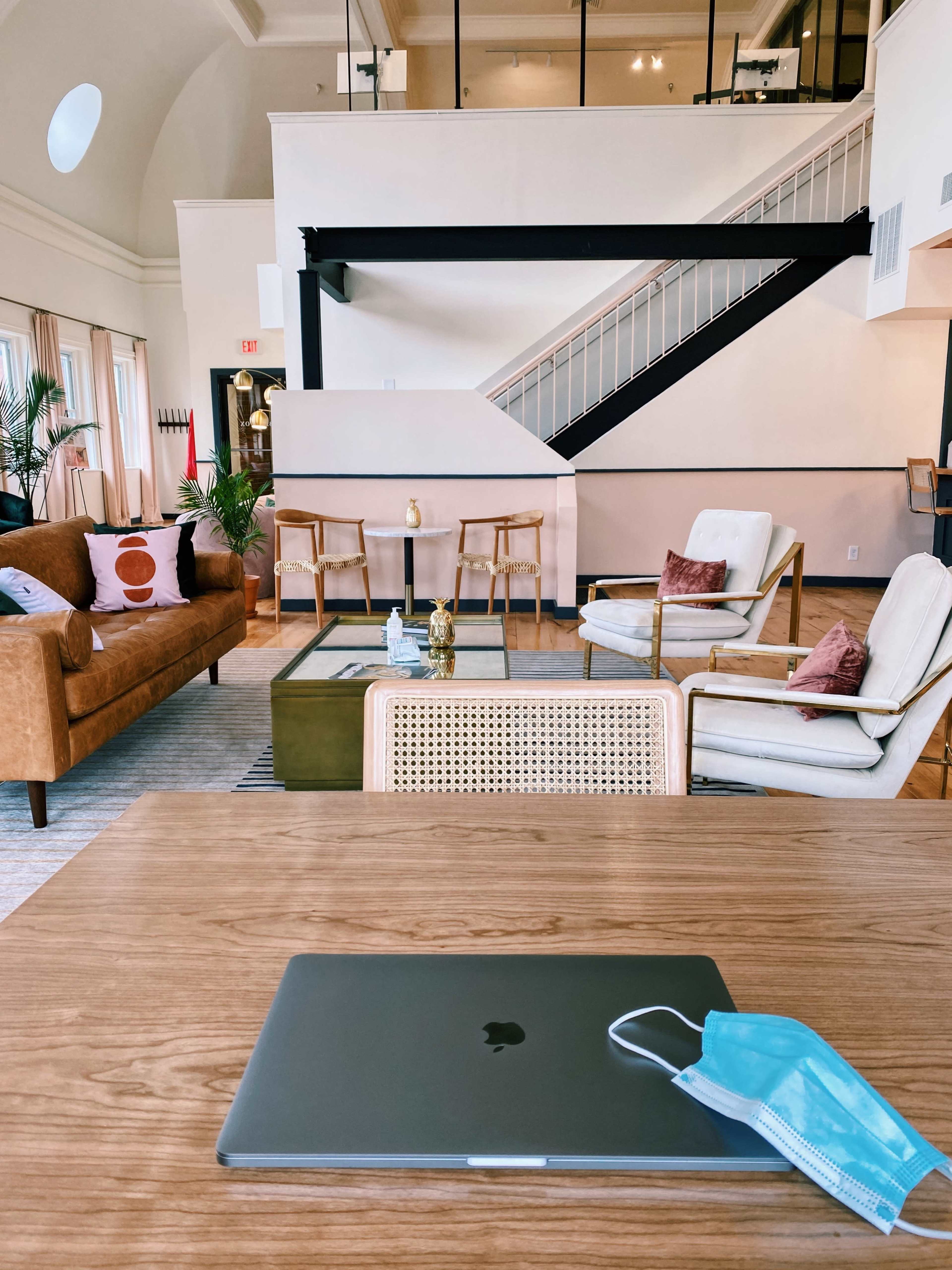 A modern, open space features a wooden table with a laptop and a blue mask in the foreground, while sofas and chairs are arranged around a central coffee table in the background.