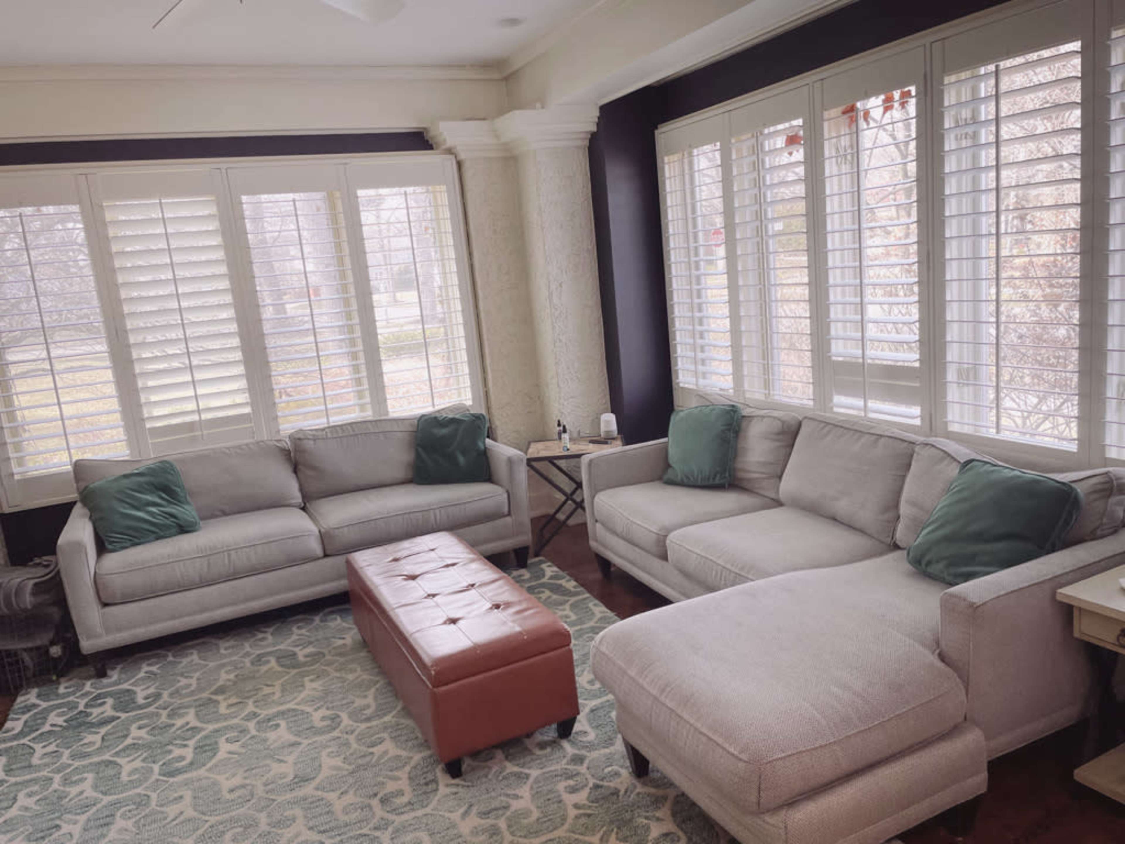 A cozy living room features two light gray sofas with green cushions, a brown ottoman, and large windows with white shutters.