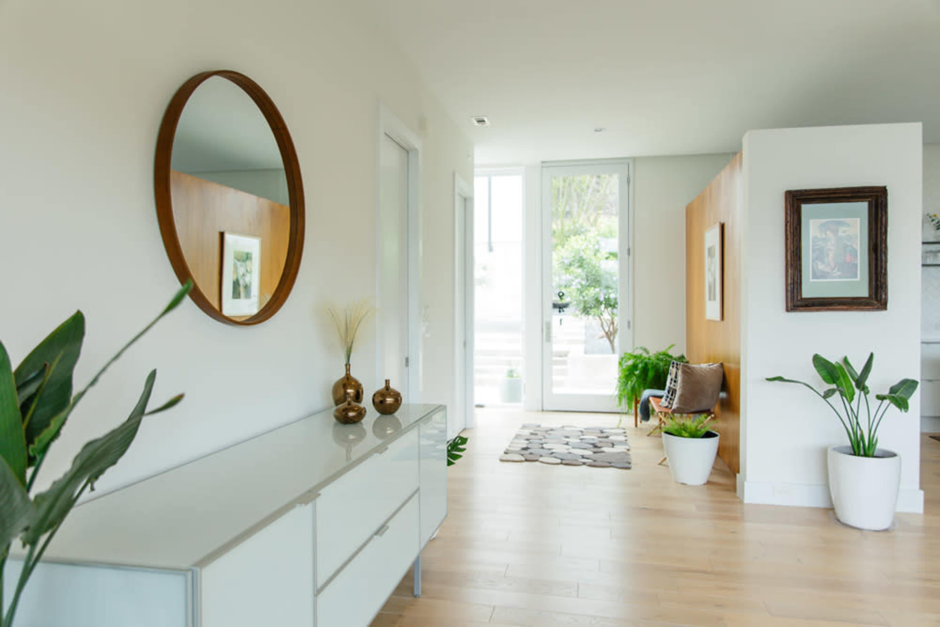 The image features a modern hallway with a round mirror, a console table, and indoor plants, leading to a doorway with natural light.