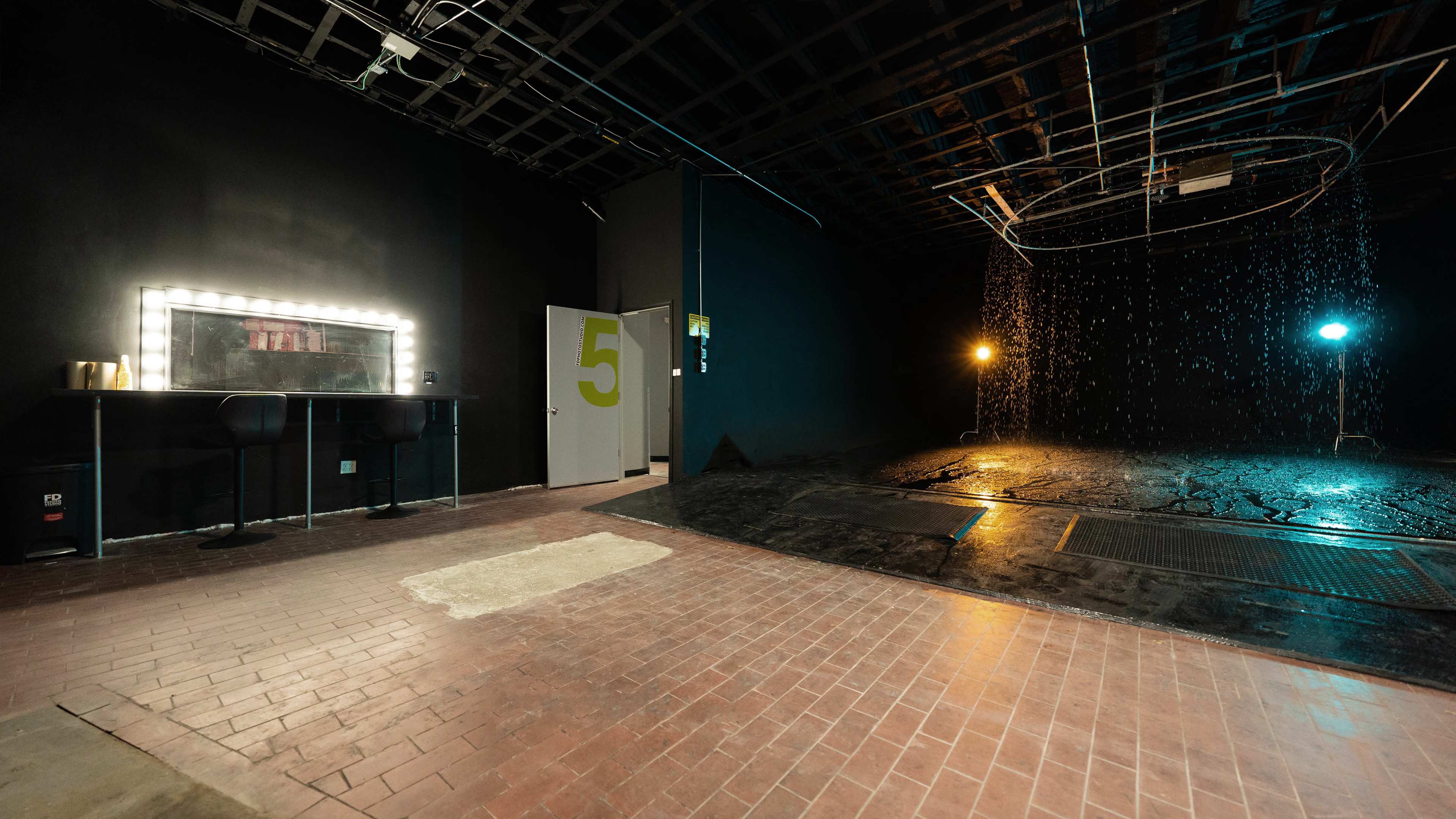 A dimly lit studio space featuring a vanity mirror, a door marked with the number five, and a section with overhead lights and a water feature.